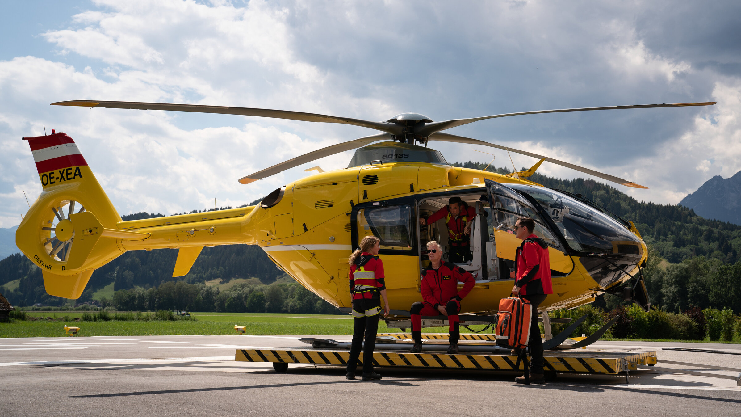 "Die Bergretter: Loslassen": Der Rettungshubschrauber steht auf dem Hanger. In der geöffneten Tür sitzt Michael (Robert Lohr), hinter ihm steht Simon (Ferdinand Seebacher). Sie blicken auf Katharina (Luise Bähr) und Rudi (Michael Pascher), die vor ihnen stehen.
