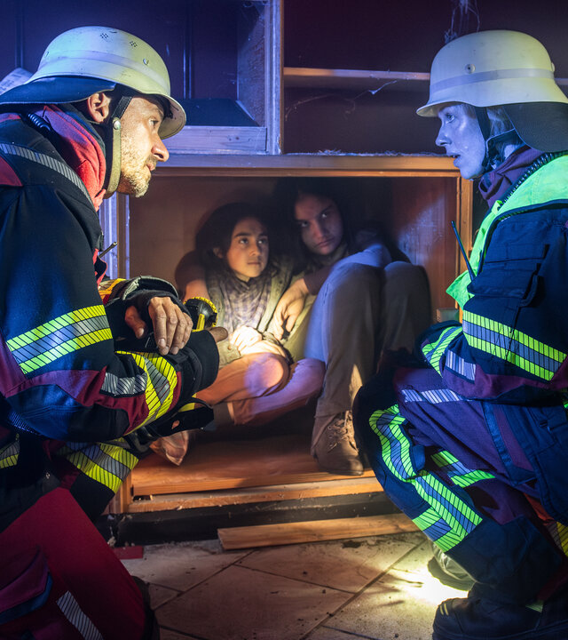 "Marie fängt Feuer - Aufbruch ins Ungewisse": Marie (Christine Eixenberger) kniet neben Peter Angerer (Stephan Luca) vor einem Holzschrank, in dem sich zwei Kinder versteckt halten. Beide tragen ihre Feuerwehruniform Helm und Taschenlampe und sehen sich an.
