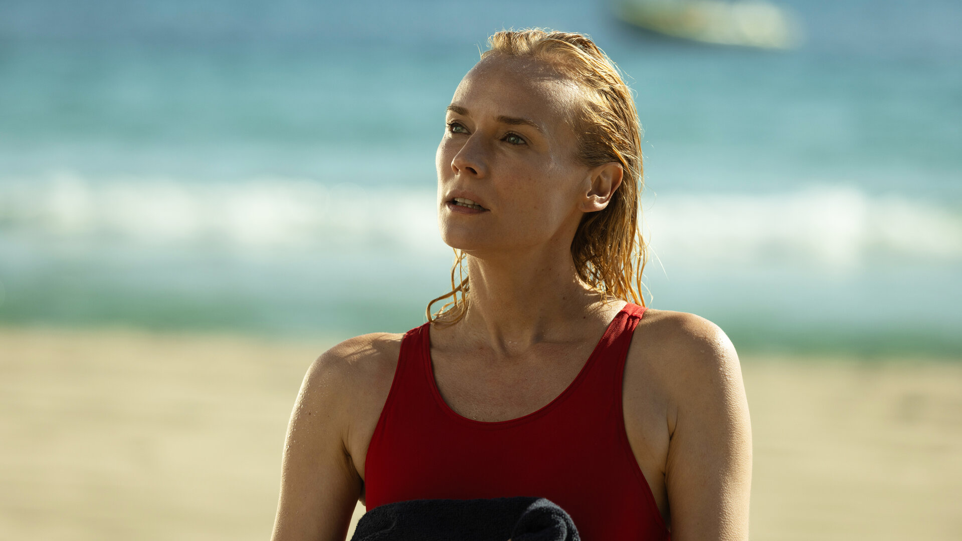 "Visions - Tödliches Verlangen": Halbnah: Estelle (Diane Kruger) steht in einem roten Badeanzug am Strand. Vor der Brust hält sie ein dunkles Handtuch, sie blickt fragend landeinwärts, im Hintergrund liegt das Meer.