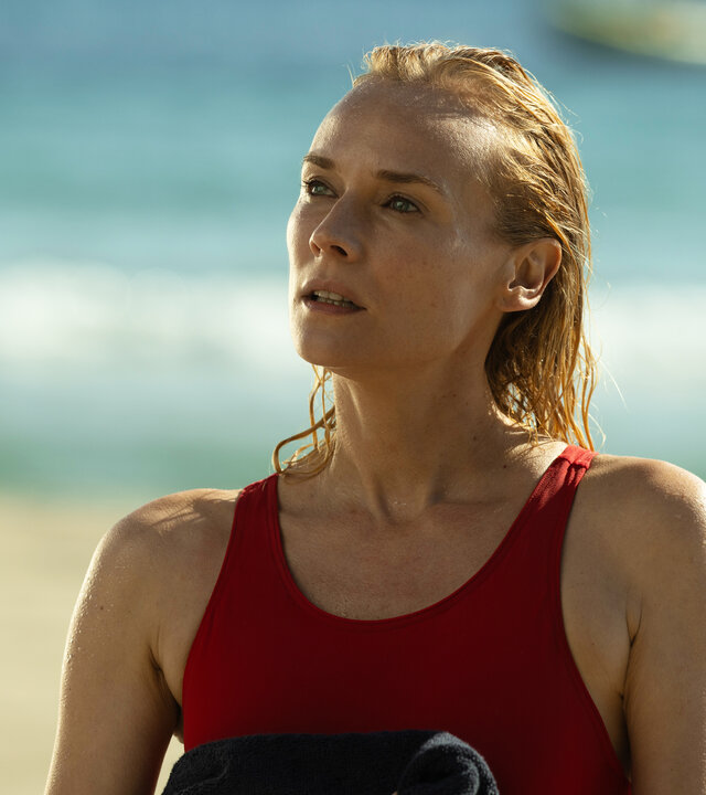 "Visions - Tödliches Verlangen": Halbnah: Estelle (Diane Kruger) steht in einem roten Badeanzug am Strand. Vor der Brust hält sie ein dunkles Handtuch, sie blickt fragend landeinwärts, im Hintergrund liegt das Meer.