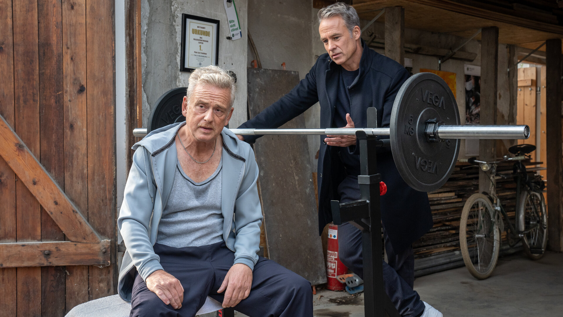 "SOKO Leipzig - Nadelstiche": Thilo Neuenburg (Thomas Huber) sitzt in einem Schuppen auf einer Hantelbank mit dem Rücken zu Jan Maybach (Marco Girnth). Die beiden Männer sprechen miteinander.