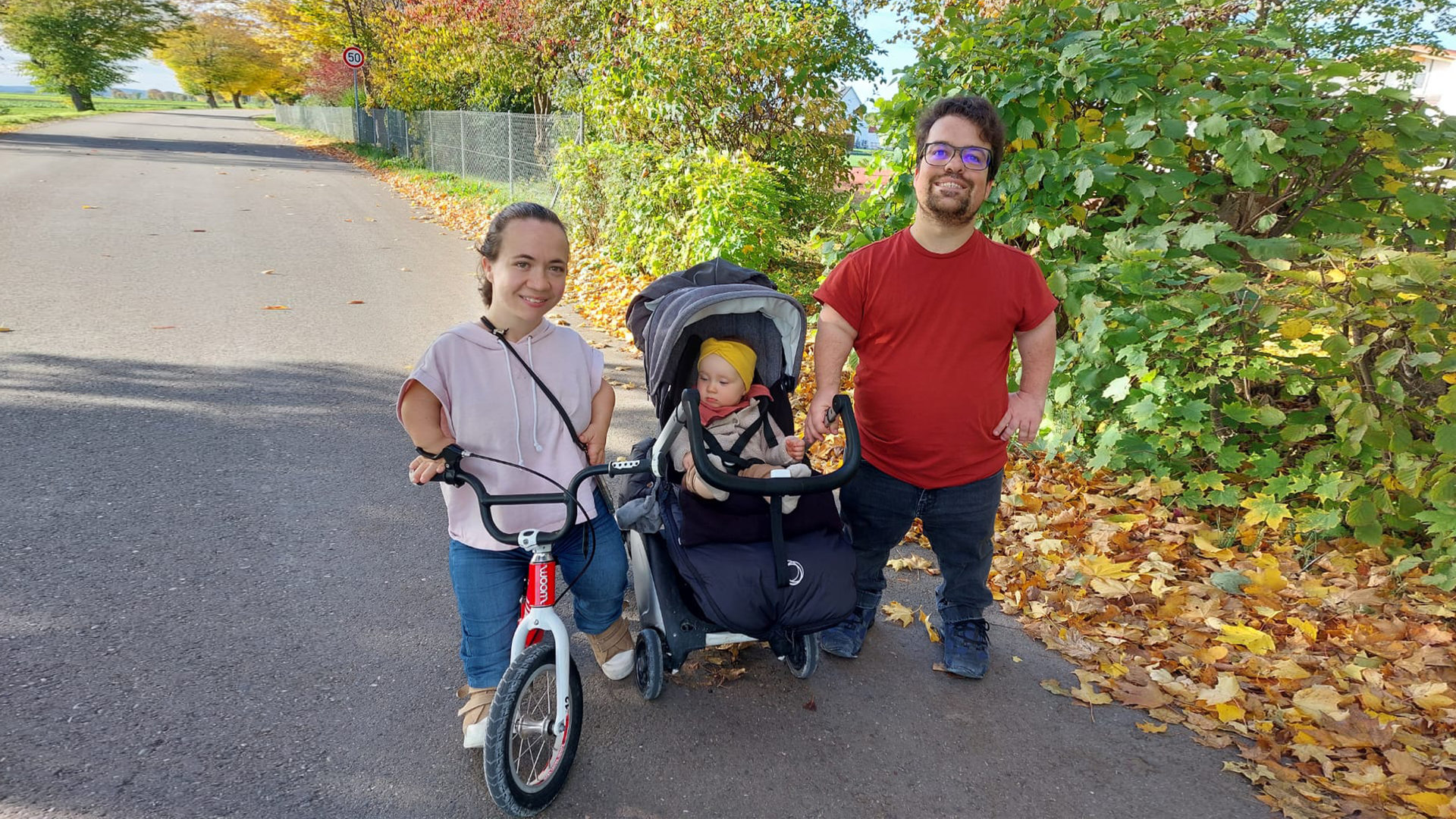 "einfach Mensch: Marlis und Gary - Eltern mit Kleinwuchs:" Die kleinwüchsigen Marlis und Gary stehen mit dem Kinderwagen auf einem Feldweg. Marlis benutzt ein Laufrad.