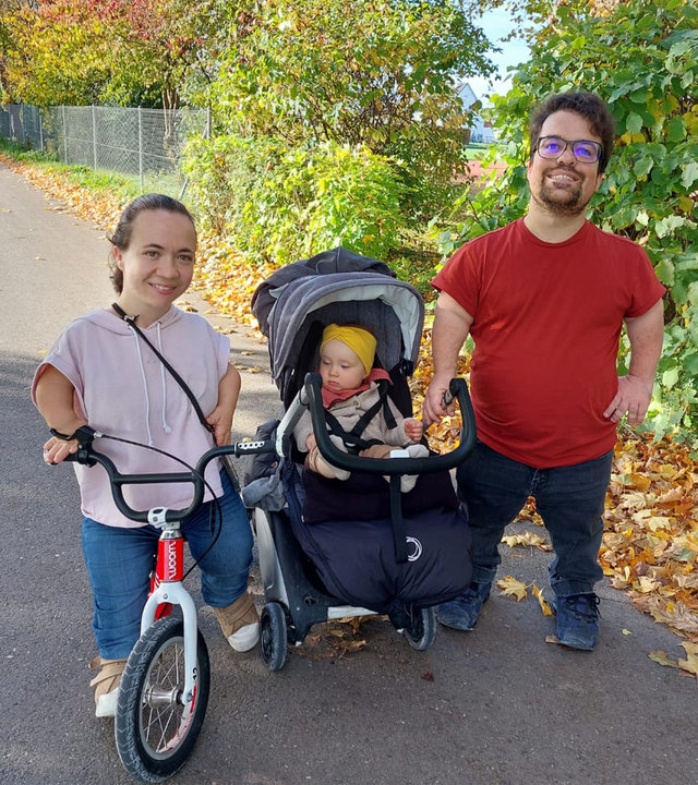 "einfach Mensch: Marlis und Gary - Eltern mit Kleinwuchs:" Die kleinwüchsigen Marlis und Gary stehen mit dem Kinderwagen auf einem Feldweg. Marlis benutzt ein Laufrad.