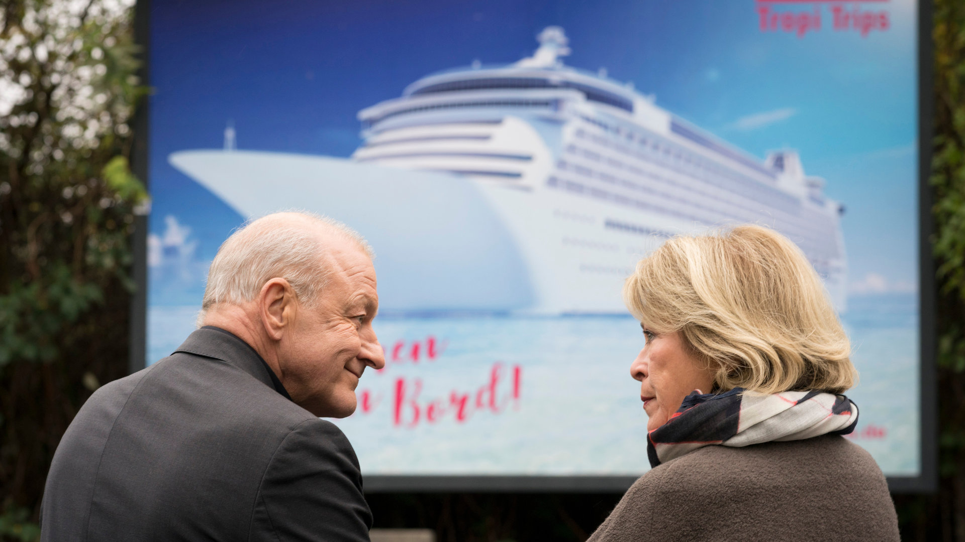 "Wilsberg - Mörderische Rendite": Georg Wilsberg (Leonard Lansink) und Anna Springer (Rita Russek) sitzen gemeinsam auf einer Bank vor einem Kreuzfahrt-Werbeplakat und schauen sich an.