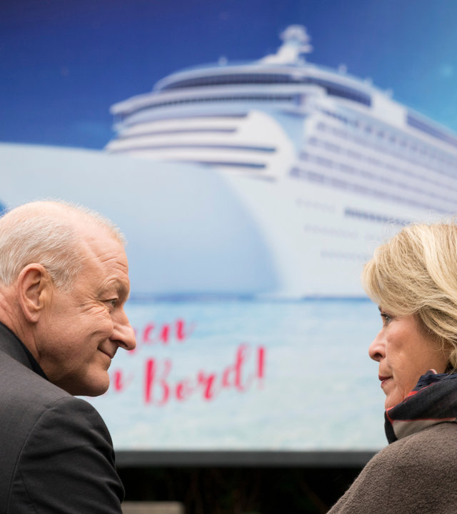 "Wilsberg - Mörderische Rendite": Georg Wilsberg (Leonard Lansink) und Anna Springer (Rita Russek) sitzen gemeinsam auf einer Bank vor einem Kreuzfahrt-Werbeplakat und schauen sich an.