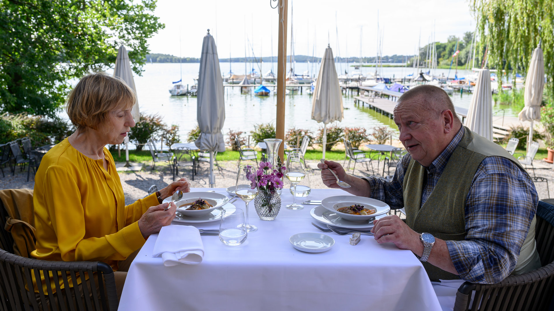 "SOKO Wismar – Der König ist tot": Dr. Helene Sturbeck (Katharina Blaschke) und Jan-Hinrich Reuter (Udo Kroschwald) essen im Restaurant "Kombüse". Sie sitzen im Freien mit Blick auf einen Bootsanleger.