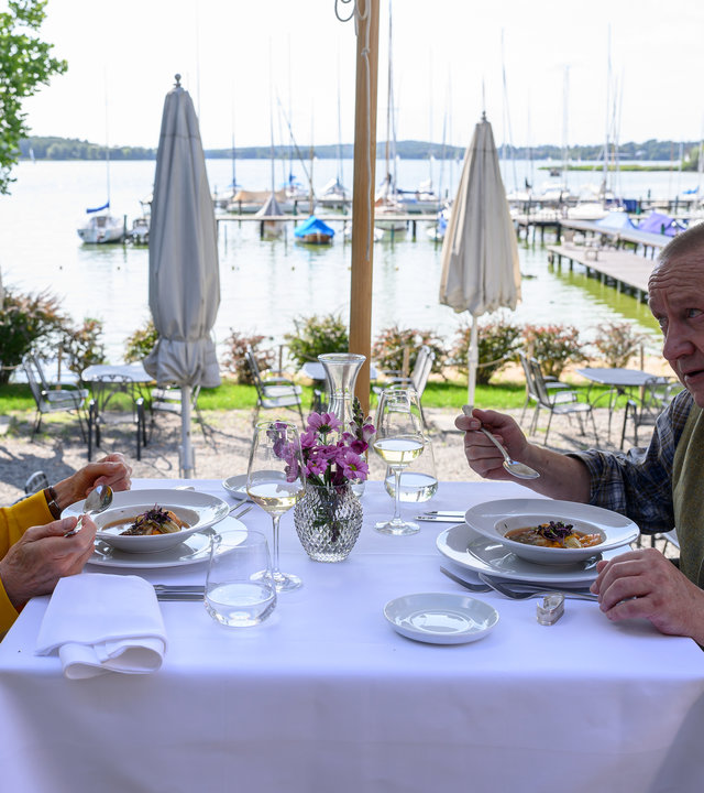 "SOKO Wismar – Der König ist tot": Dr. Helene Sturbeck (Katharina Blaschke) und Jan-Hinrich Reuter (Udo Kroschwald) essen im Restaurant "Kombüse". Sie sitzen im Freien mit Blick auf einen Bootsanleger.