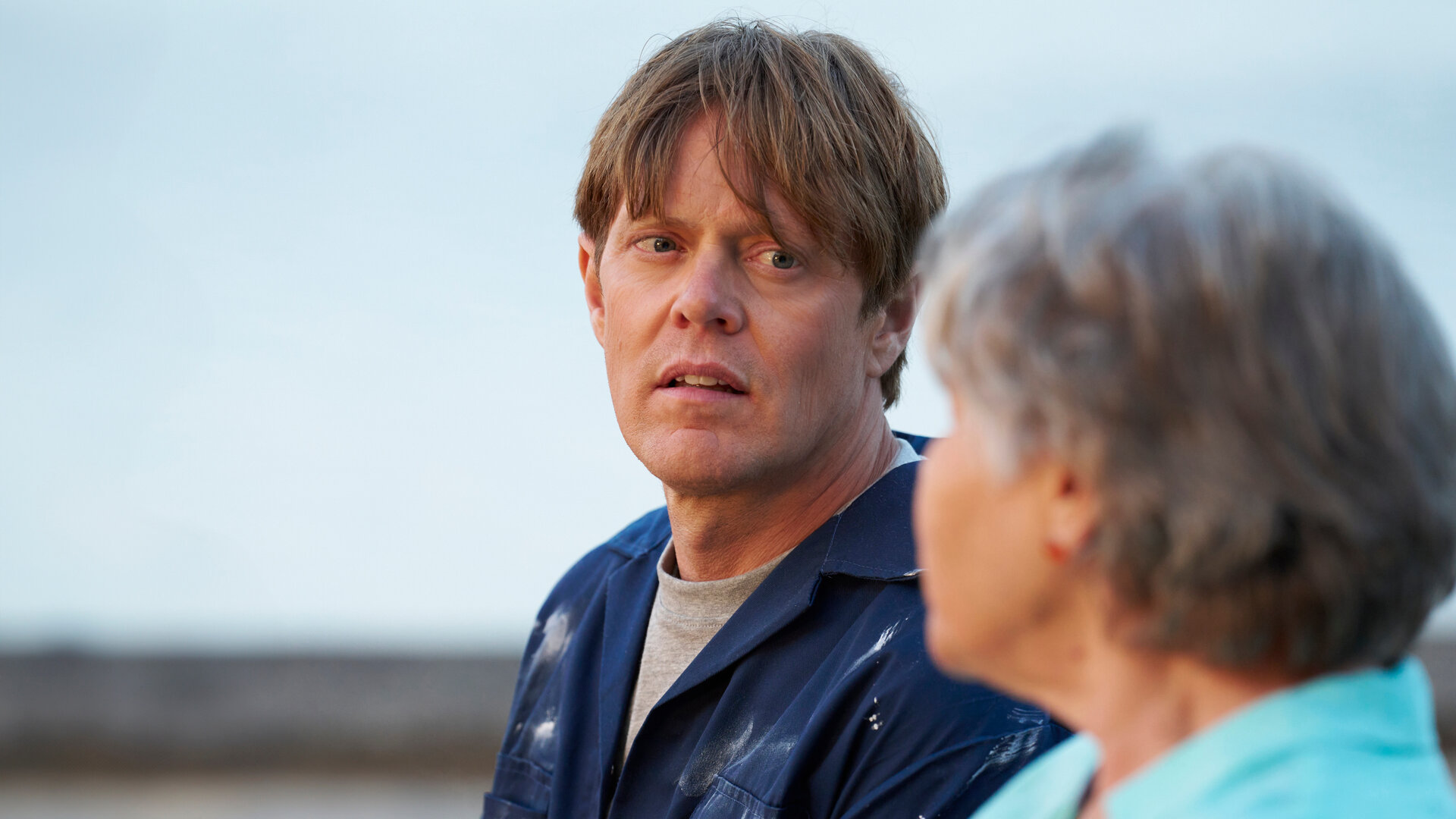 "Beyond Paradise - Die Vorhersage": Humphrey Goodman (Kris Marshall) sitzt neben Anne Lloyd (Barbara Flynn), beide haben sich einander zugewandt. Anne ist unscharf im Vordergrund und nur im Profil zu sehen. Humphrey trägt einen farbverschmierten blauen Overall.
