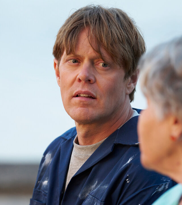 "Beyond Paradise - Die Vorhersage": Humphrey Goodman (Kris Marshall) sitzt neben Anne Lloyd (Barbara Flynn), beide haben sich einander zugewandt. Anne ist unscharf im Vordergrund und nur im Profil zu sehen. Humphrey trägt einen farbverschmierten blauen Overall.