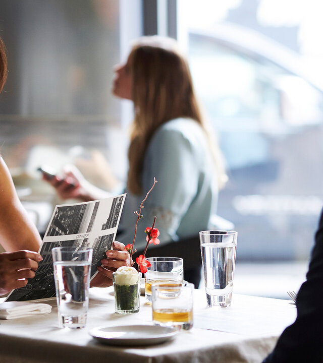 "Suits - Teile und herrsche": Jessica (Gina Torres), in weiß gekleidet, sitzt Harvey (Gabriel Macht) in einem Restaurant gegenüber, dessen Rücken der Kamera zugewandt ist. Während sie ihn anschaut, hält sie mit beiden Händen ein Papier. Im Hintergrund ist eine Frau an einem anderen Tisch zu sehen.