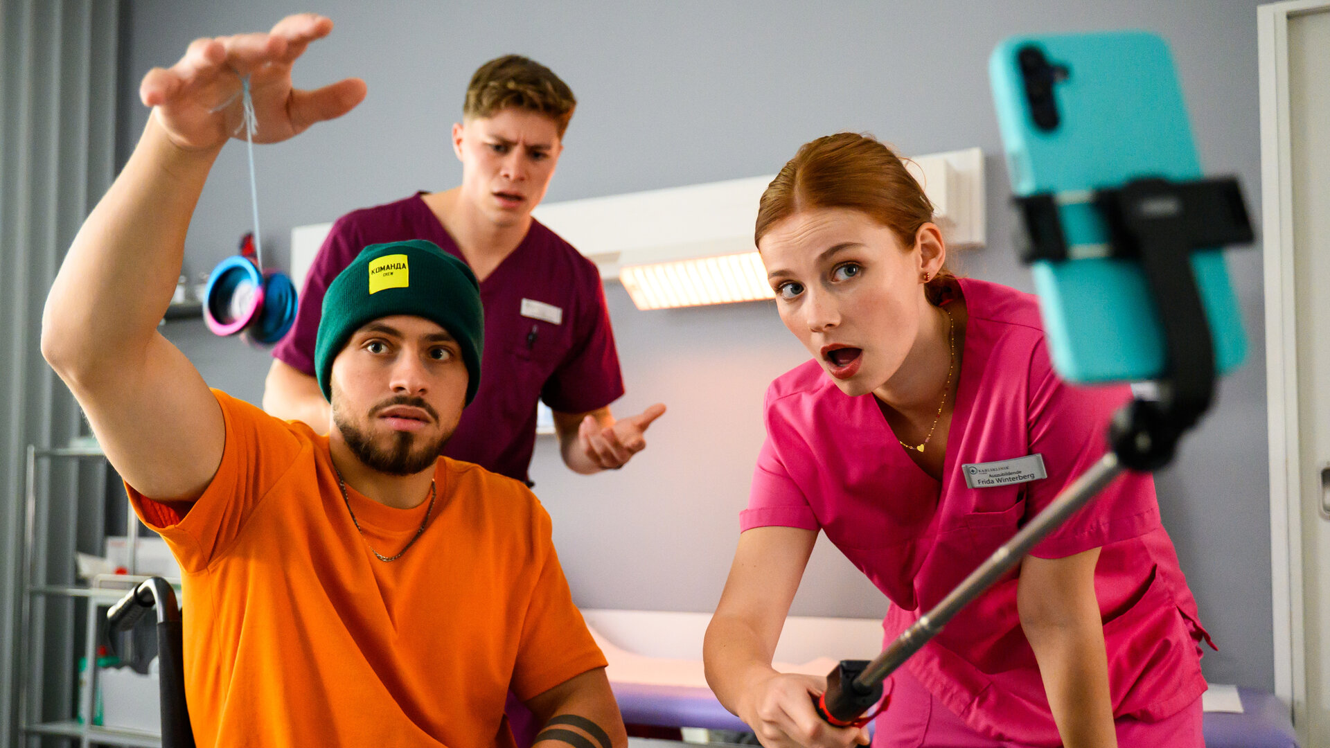"Bettys Diagnose - Große Kinder, große Sorgen": In einem Raum in der Klinik: Amir (Angelo Alabiso) hält ein Jo-Jo in der Hand und wird dabei von Frida (Johanna Hens) mit dem Handy gefilmt. Sie hält dazu einen Selfiestick in seine Richtung. Ivo (Oscar Nadermann) steht im Hintergrund und beobachtet das Geschehen.