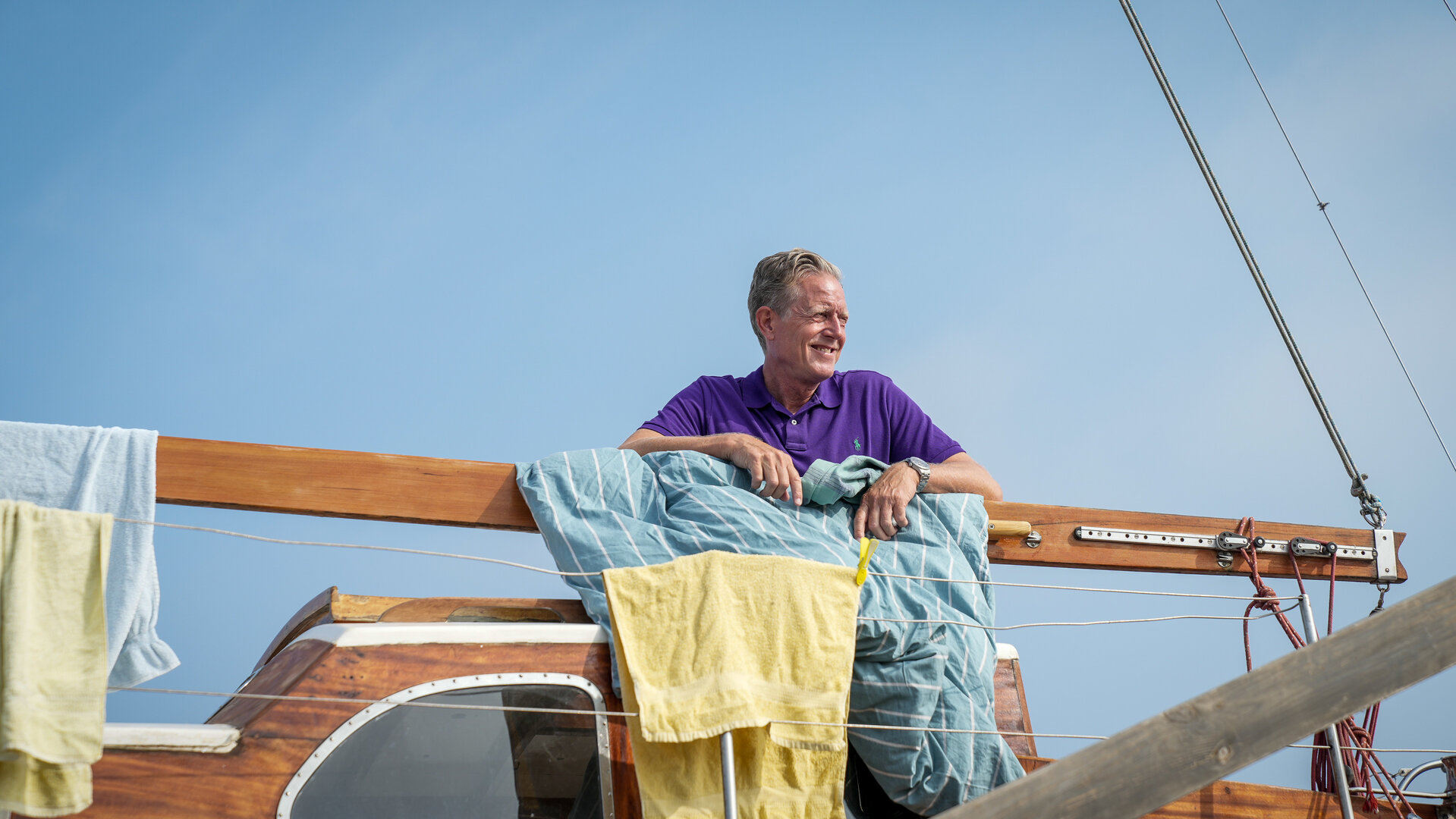 "Dan Sommerdahl – Tödliche Idylle - Wo ist Belle Helene": Dan (Peter Mygind) steht lächelnd auf dem Deck seines Bootes, über dem Geländer hängen Handtücher und Bettwäsche.
