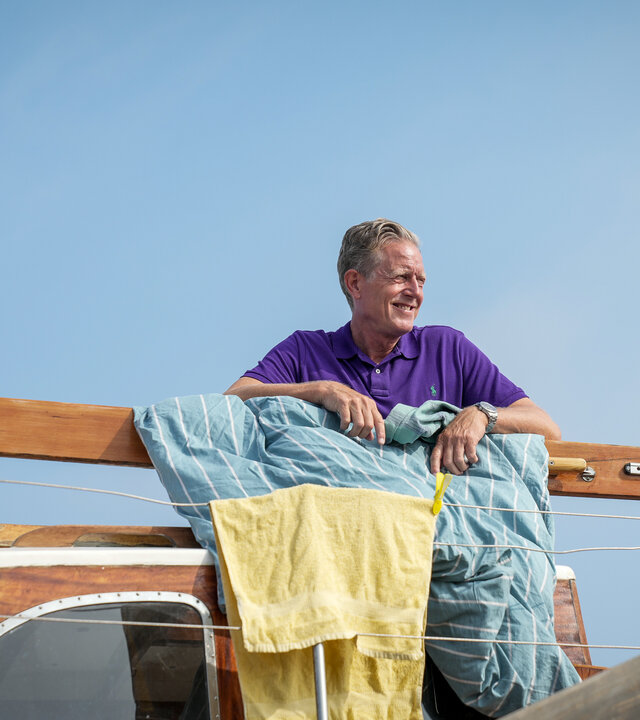 "Dan Sommerdahl – Tödliche Idylle - Wo ist Belle Helene": Dan (Peter Mygind) steht lächelnd auf dem Deck seines Bootes, über dem Geländer hängen Handtücher und Bettwäsche.
