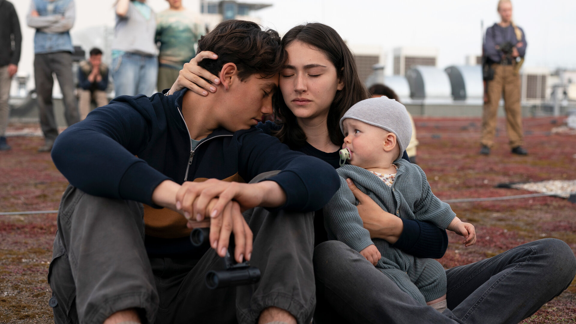 "Sløborn III – Eine neue Welt": Alex (Ron Renzenbrink) und Evelin (Emily Kusche) sitzen eng umschlungen mit Baby Mila (Lucia Wernze) im Arm auf einem mit Moos bewachsenen Flachdach. Im Hintergrund stehen mehrere Menschen.