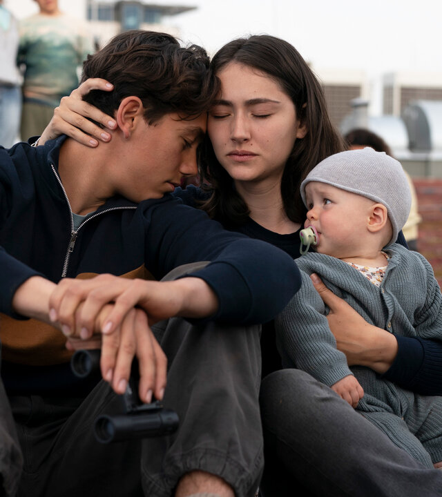"Sløborn III – Eine neue Welt": Alex (Ron Renzenbrink) und Evelin (Emily Kusche) sitzen eng umschlungen mit Baby Mila (Lucia Wernze) im Arm auf einem mit Moos bewachsenen Flachdach. Im Hintergrund stehen mehrere Menschen.