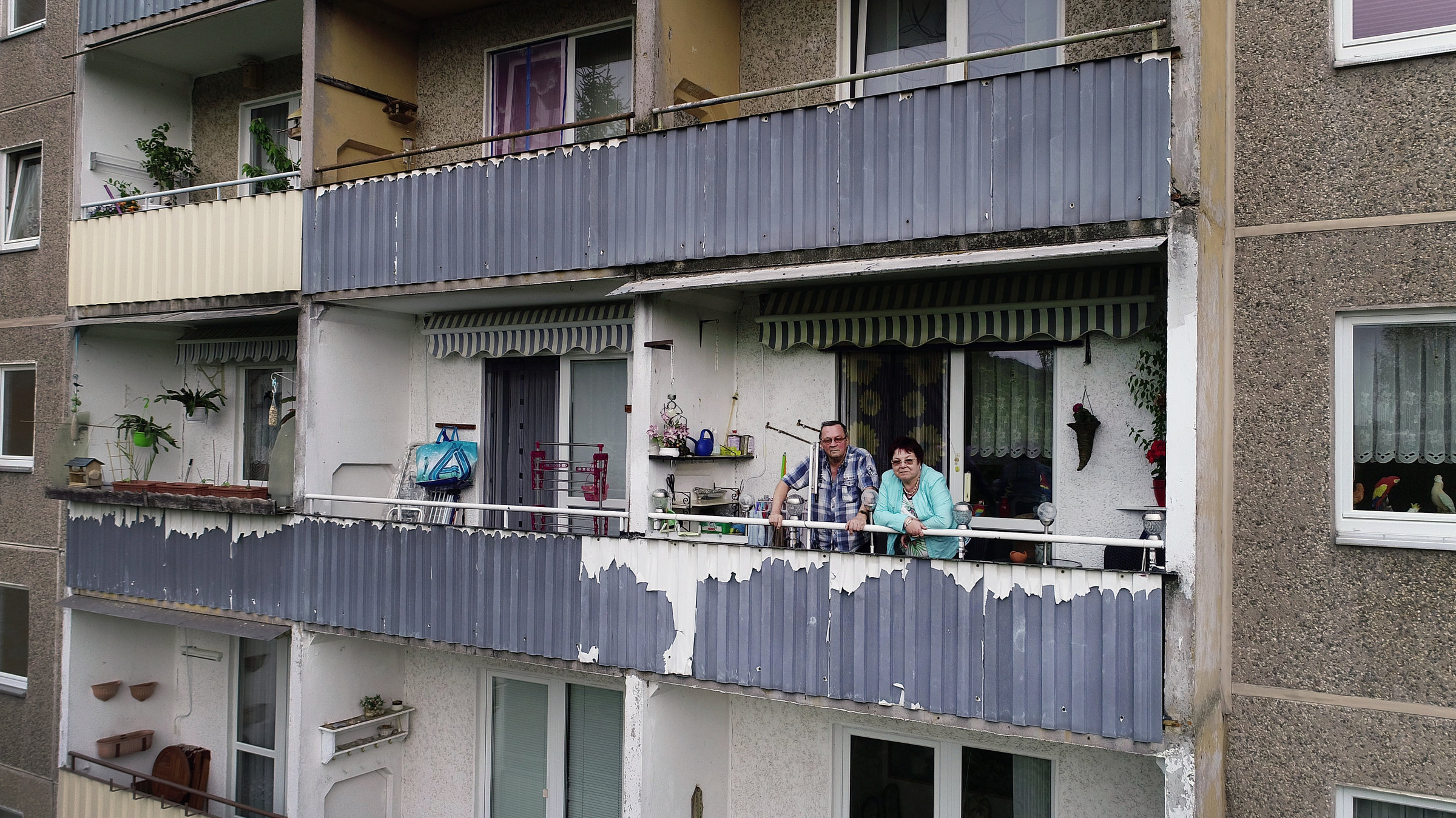 "37°: Plattgemacht - Wenn ein Stadtteil verschwindet": Christine und Hans stehen auf einem Balkon einem Plattenbau. Das Gebäude wirkt verfallen, die Farbe blättert von der Fassade.