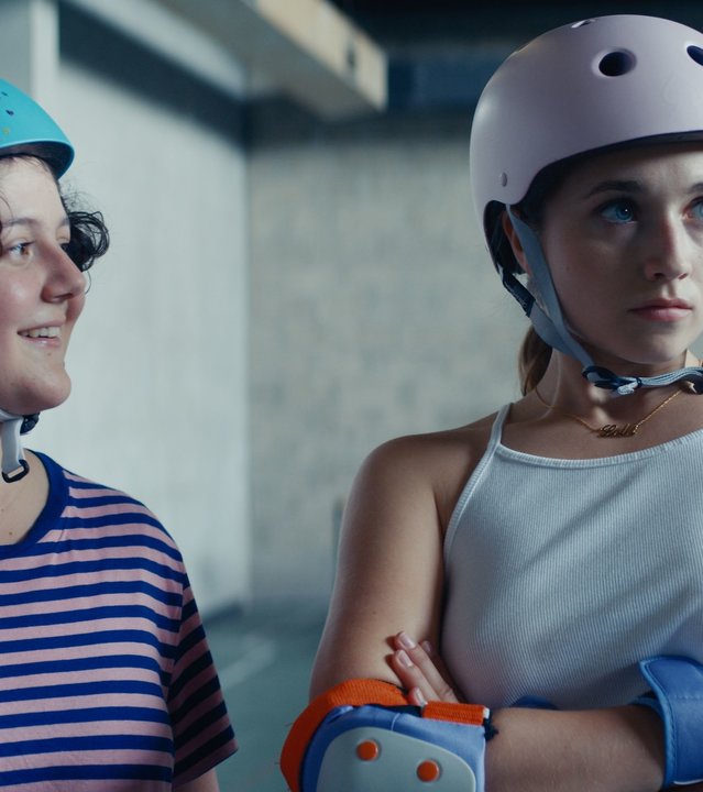 "Derby Girl - Lektionen": Lola Bouvier (Chloé Jouannet) hat beide Arme vor ihrer Brust verschränkt und schaut genervt in die Ferne. Marie-Josée (Salomé Diénis Meulien), die sich MJ nennt, steht neben Lola und blickt sie lächelnd von der Seite an. Beide tragen Sportklamotten und einen Helm.