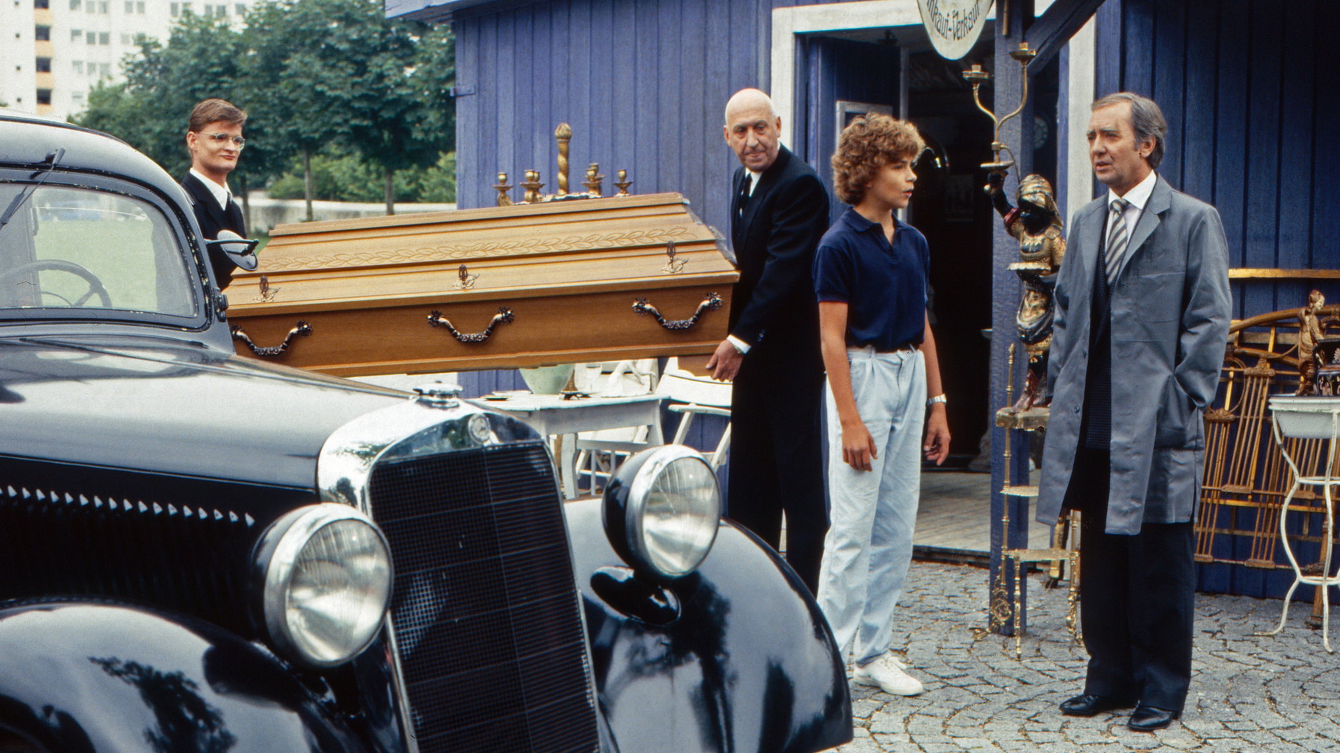 "Oliver Maass - Das Spiel mit der Zaubergeige": Oliver (Josef Gröbmayr) und Esteban (Hans Clarin) unterhalten sich, während Eickelberg (Karl Lieffen) mit einem unbekannten Kollegen einen Sarg zu einem Auto trägt.