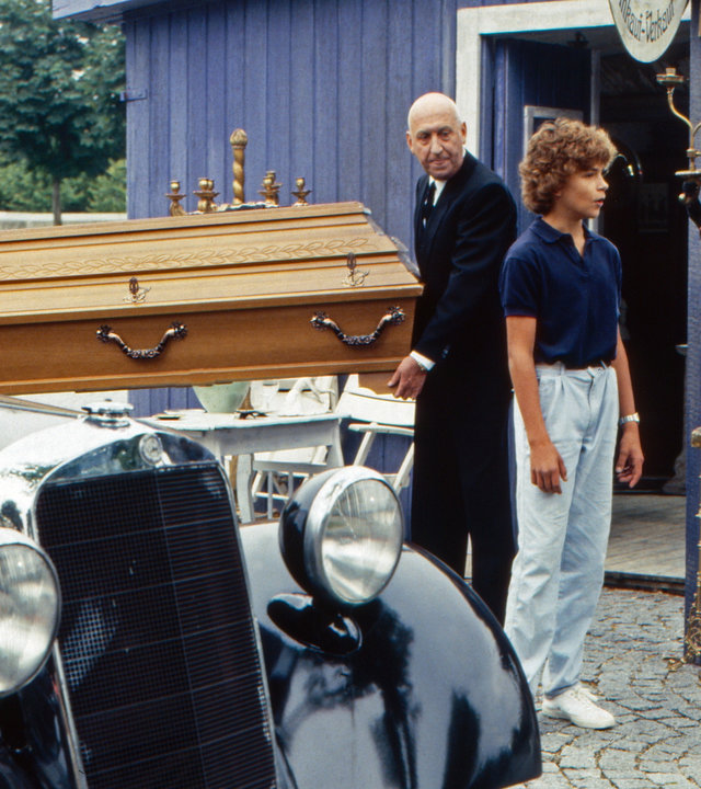 "Oliver Maass - Das Spiel mit der Zaubergeige": Oliver (Josef Gröbmayr) und Esteban (Hans Clarin) unterhalten sich, während Eickelberg (Karl Lieffen) mit einem unbekannten Kollegen einen Sarg zu einem Auto trägt.