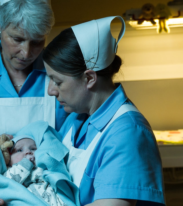 "SOKO Stuttgart - Babyklappe": Schwester Evi Neumann (Tilla Kratochwil) hält ein Neugeborenes (Komparse) in den Armen, während eine weitere Schwester (Komparsin) ihr assistiert. Beide tragen hellblaue Schwesterntracht mit weißen Schürze und weißen Häubchen. Sie stehen vor der geöffneten Babyklappe.