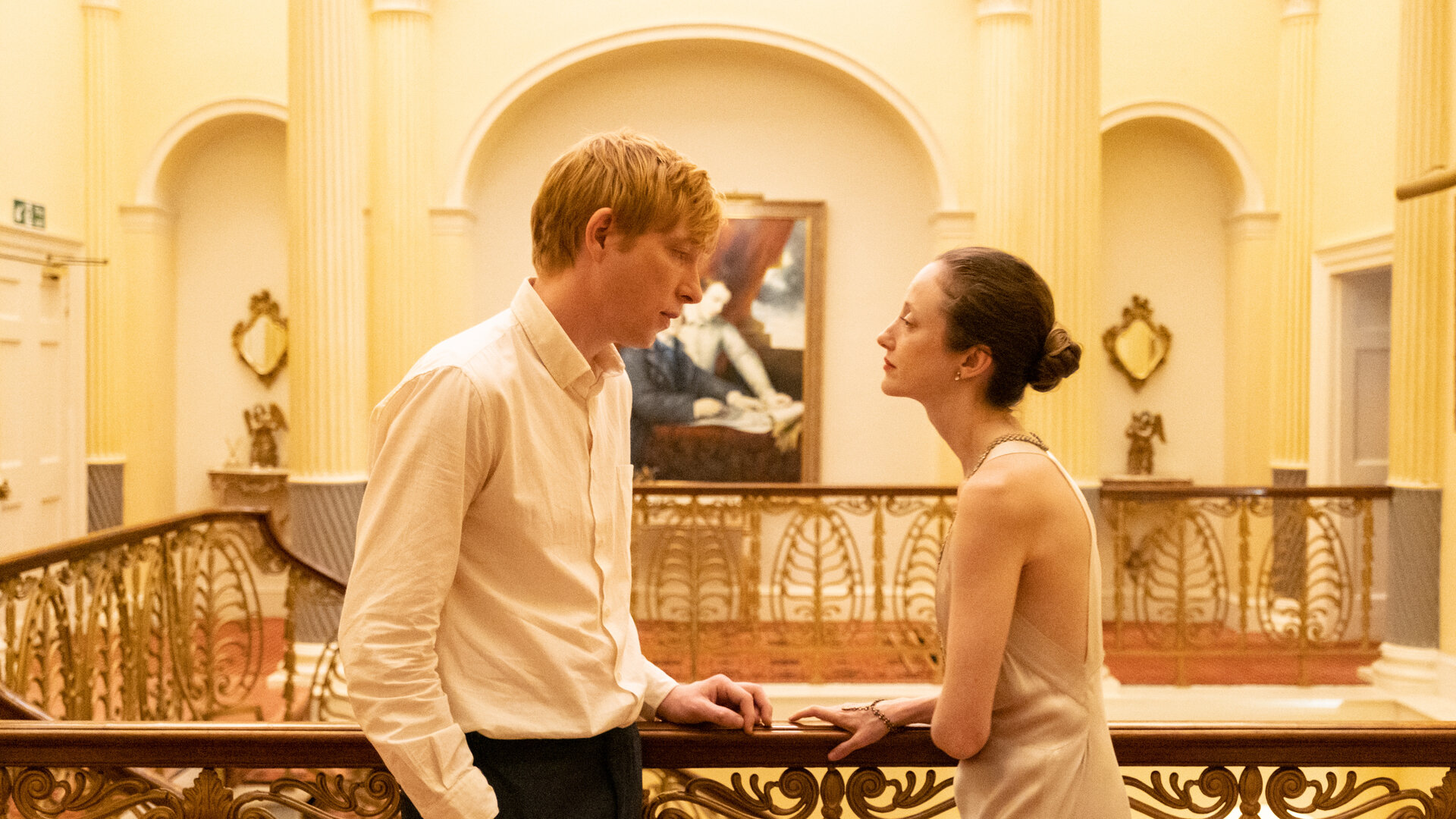 "Alice & Jack - Risiko": Jack (Domhnall Glesson) und Alice (Andrea Riseborouht) stehen sich Innen am Treppengeländer eines wunderschönen Hauses gegenüber. Er steht lässig, mit den Händen in den Taschen da, während sie die Arme schützend verschränkt vor sich hält.