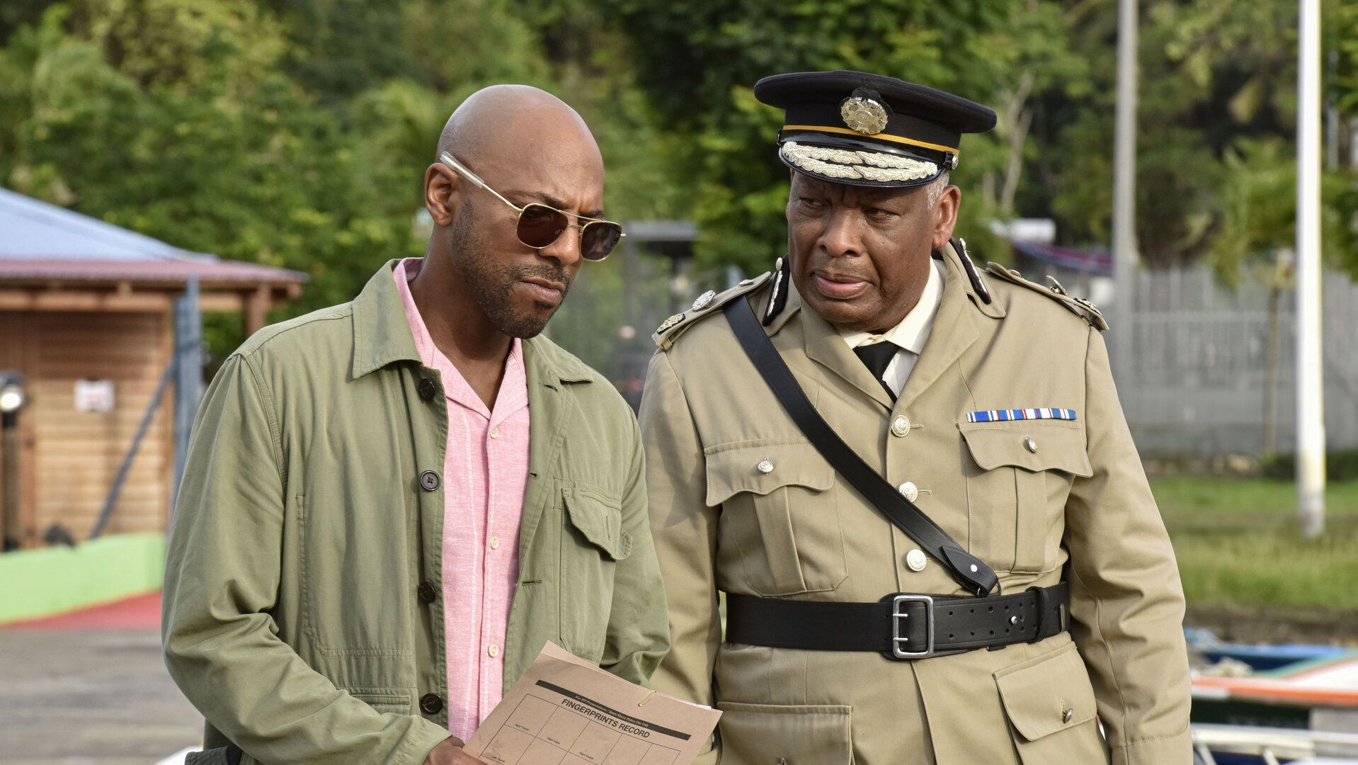 "Death in Paradise - Maria": Detective Mervin Wilson (Don Gilet) und Commissioner Selwyn Patterson (Don Warrington) stehen nebeneinander auf dem Steg eines kleinen Hafens. Mervin trägt eine Sonnenbrille und hält ein großes Blatt Papier in den Händen. Sein Blick ist fragend nach vorn gerichtet. Selwyn trägt seine helle Dienstuniform sowie die dazugehörige schwarze Dienstmütze.