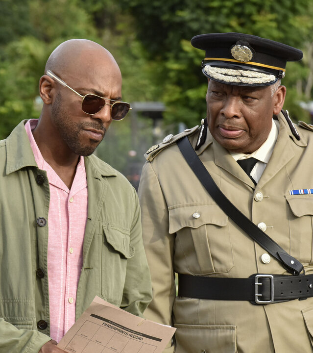 "Death in Paradise - Maria": Detective Mervin Wilson (Don Gilet) und Commissioner Selwyn Patterson (Don Warrington) stehen nebeneinander auf dem Steg eines kleinen Hafens. Mervin trägt eine Sonnenbrille und hält ein großes Blatt Papier in den Händen. Sein Blick ist fragend nach vorn gerichtet. Selwyn trägt seine helle Dienstuniform sowie die dazugehörige schwarze Dienstmütze.