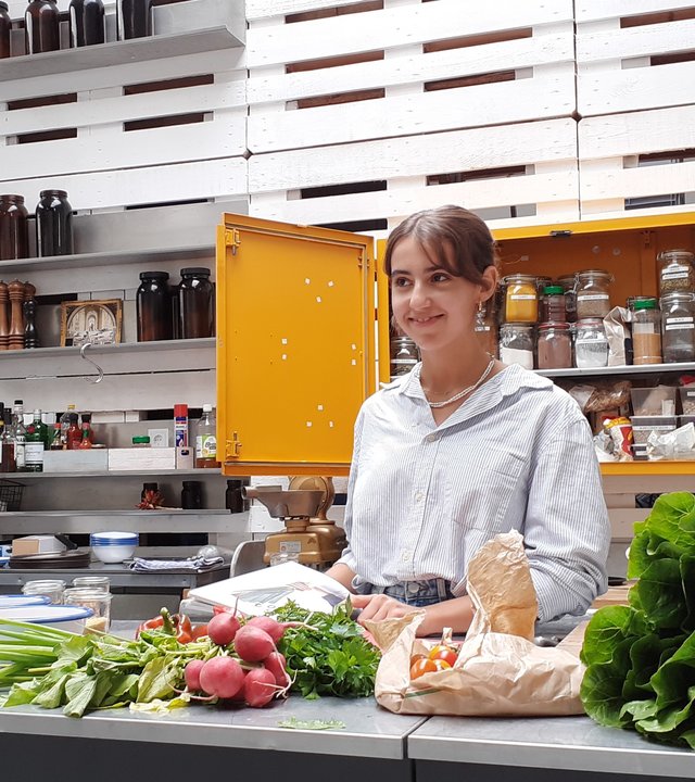 "Terra Xpress: Gesucht - das gesunde Essen": Lea Elci steht in vor einer Küche. Vor ihr liegt frisches Gemüse auf der Küchenarbeitsplatte. Hinter ihr ist ein Schrank geöffnet, in diesem stehen diverse, gefüllte Gläser mit zum Beispiel Gewürzen.