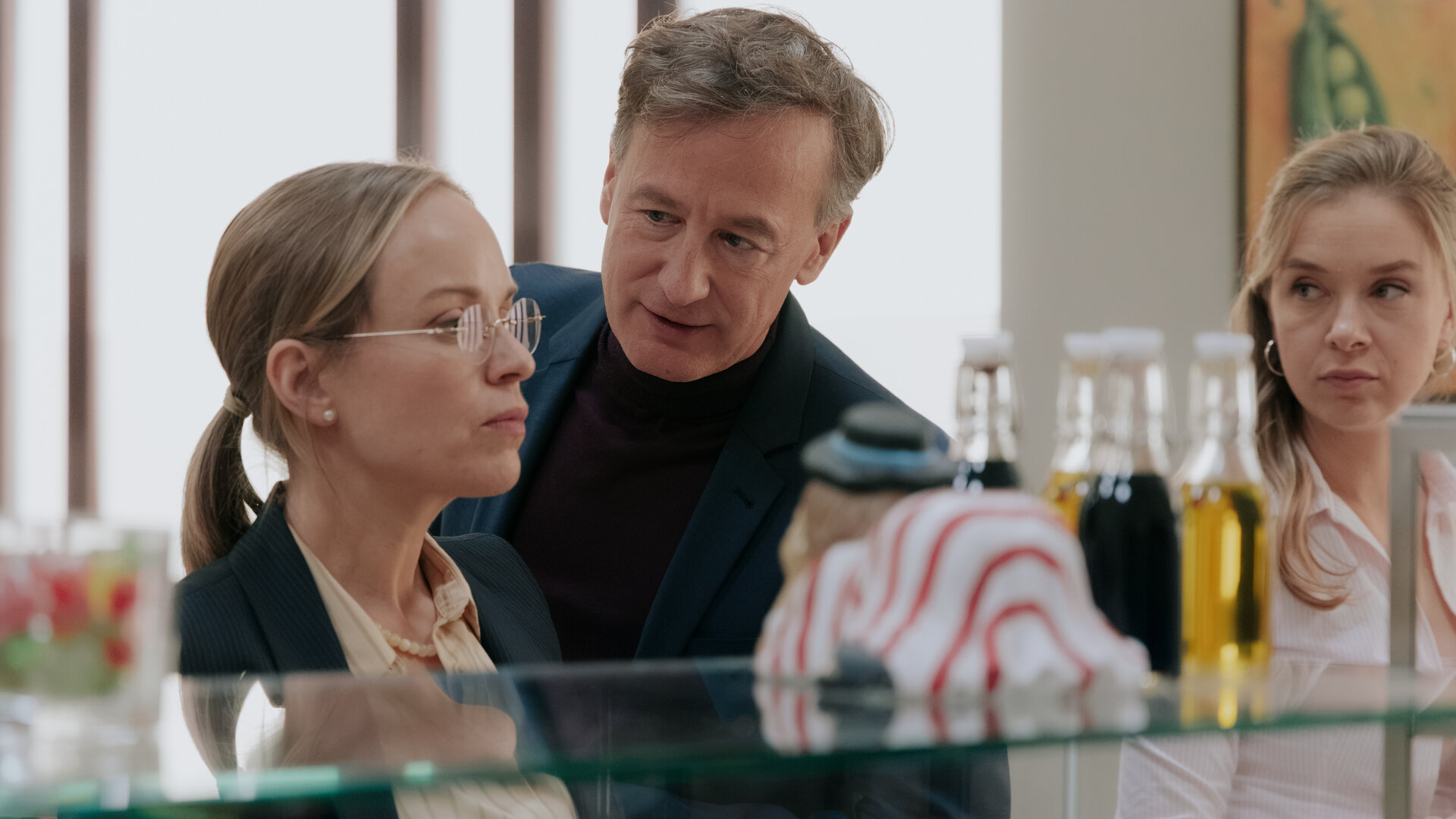 "Nine to Five - Das Amt - Tag 1": Ronja Keller (Friederike Kempter), Amtsleiter Schulenberg (Jan Henrik Stahlberg) und Melissa Seibel (Anna Herrmann) stehen nebeneinander vor der Theke in der Kantine. Georg blickt zu Ronja, Ronja blickt in die Ferne. Melissa beobachtet die beiden.