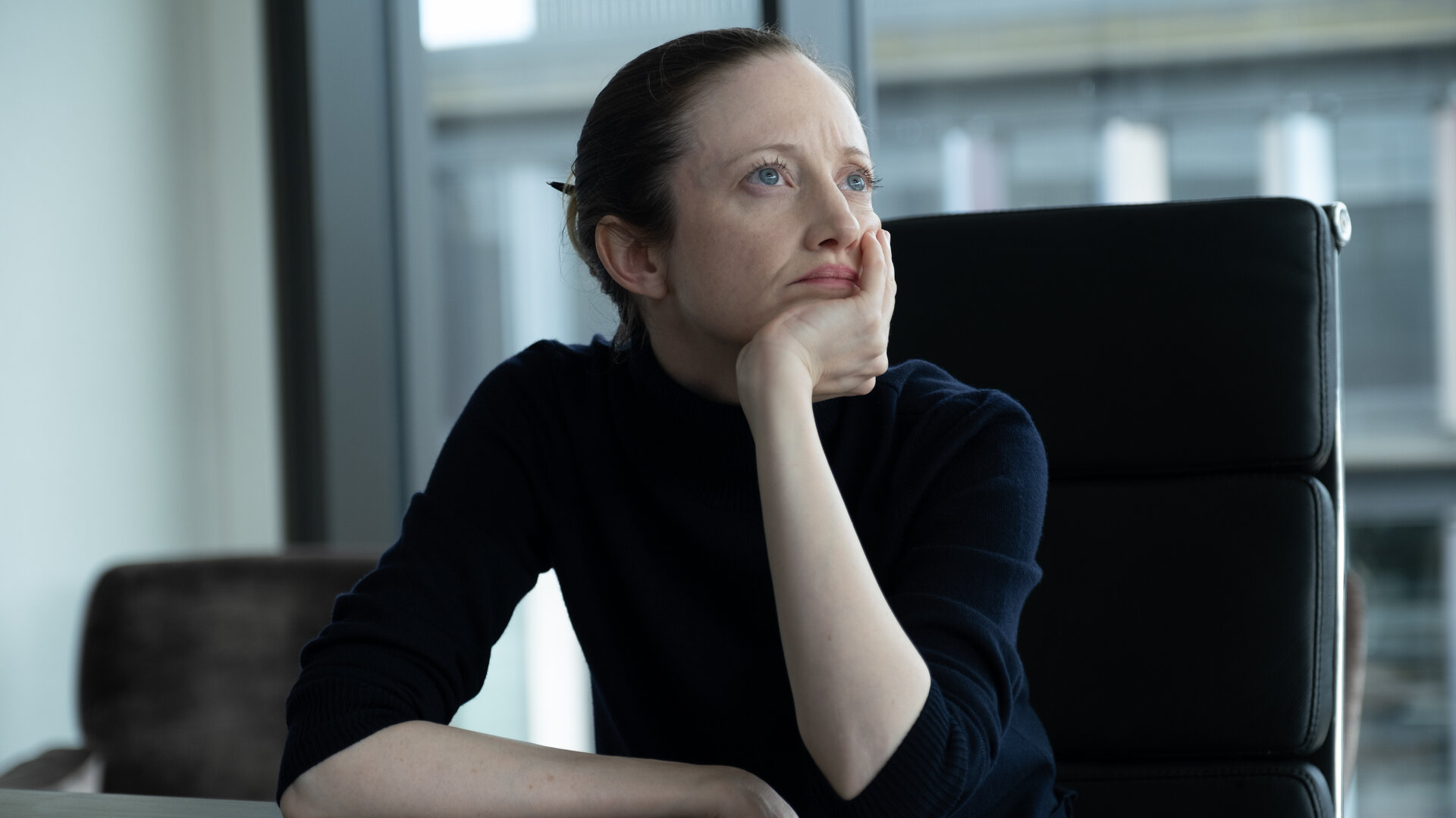 "Alice & Jack - Freundschaft": Alice (Andrea Riseborough) sitzt in einem großen schwarzen Bürostuhl an ihrem Schreibtisch, den Kopf auf die Hand gestüzt und wirkt nachdenklich. Hinter ihr eröffnet ein großes Bürofenster den Blick auf ein anderes Gebäude.