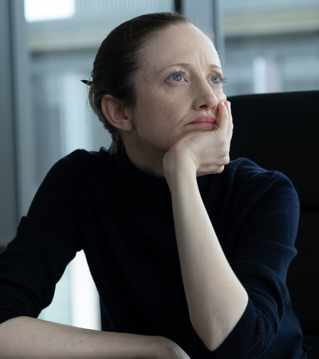 "Alice & Jack - Freundschaft": Alice (Andrea Riseborough) sitzt in einem großen schwarzen Bürostuhl an ihrem Schreibtisch, den Kopf auf die Hand gestüzt und wirkt nachdenklich. Hinter ihr eröffnet ein großes Bürofenster den Blick auf ein anderes Gebäude.