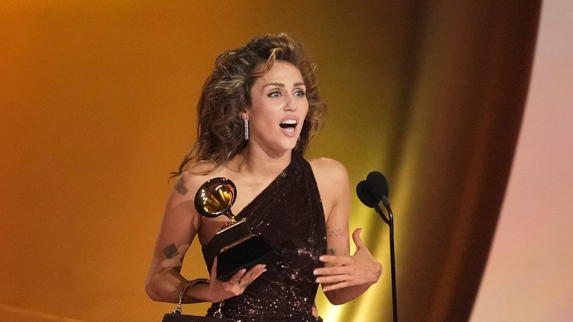 "The True Story of Miley Cyrus": Frau im Abendkleid auf Bühne vor Mikrofon, hält Auszeichnung in rechter Hand.