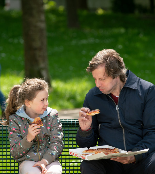 "Wilsberg: Vaterfreuden": Hanna (Charlotte Schliewe) sitzt mit Ekki Talkötter (Oliver Korittke) auf einer Parkbank. Die beiden schauen zufrieden drein und essen Pizza.
