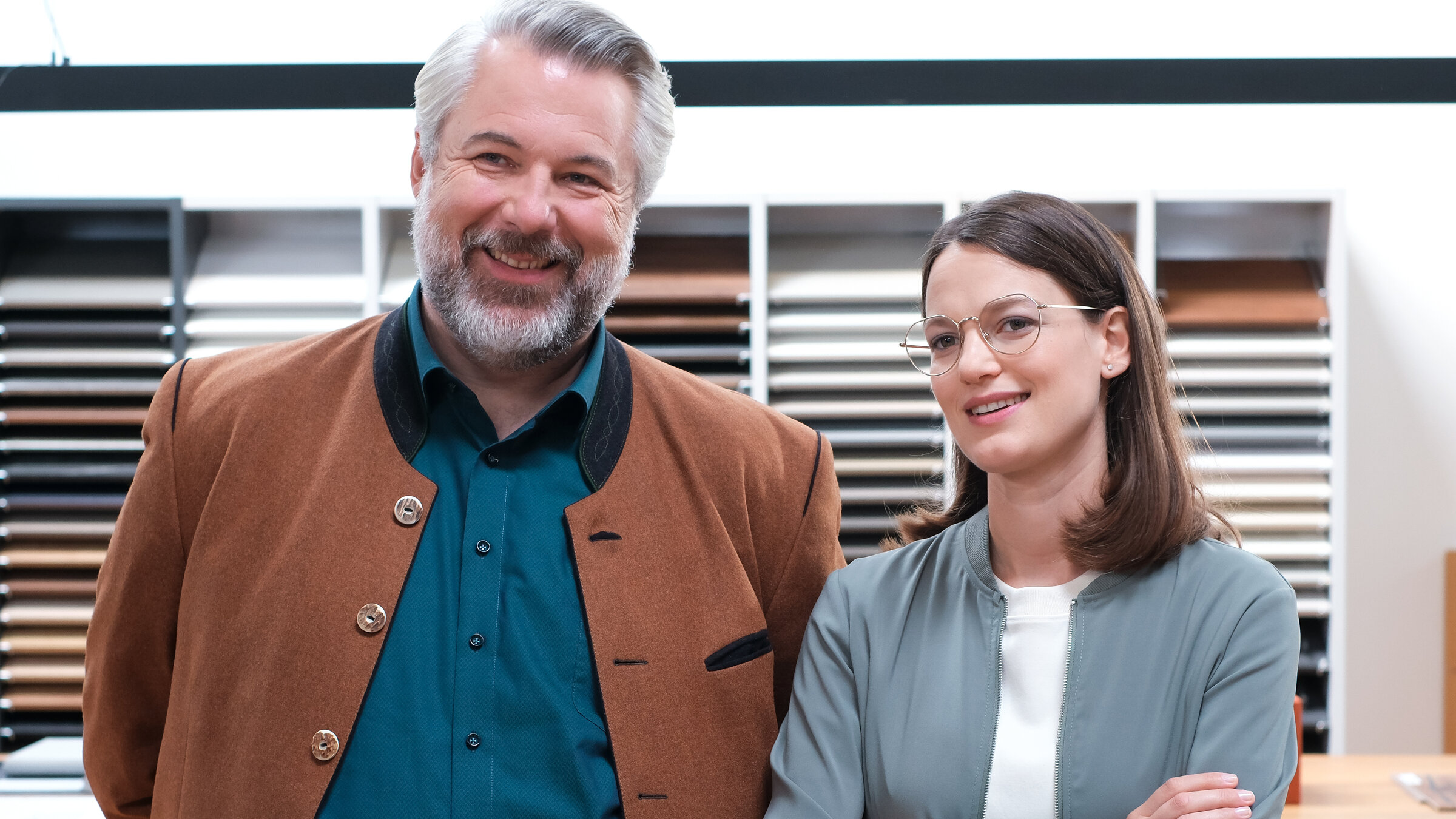 "Die Rosenheim-Cops - Eine geniale Idee": Die Kommissare Stadler (Dieter Fischer) und Beck (Michaela Weingartner) stehen lächelnd nebeneinander und schauen in dieselbe Richtung. Im Hintergrund befindet sich ein Regal mit Holztafeln.