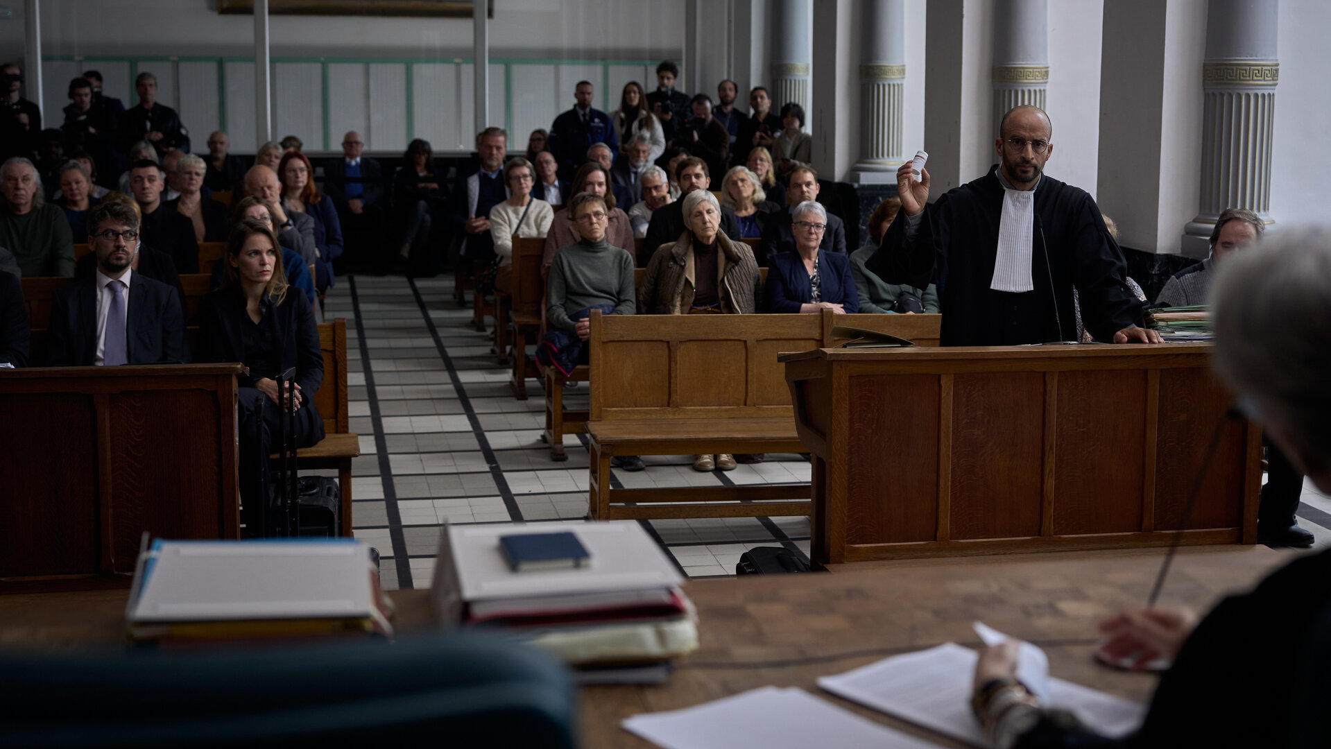 "Elixir – The Pain Killers - Deals": Hamza Toumi (Walid Benmbarek) steht in einem voll besetzten Gerichtssaal hinter dem Anwaltspult und gestikuliert. Im Vordergrund ist die Richterin von hinten im Anschnitt zu sehen.