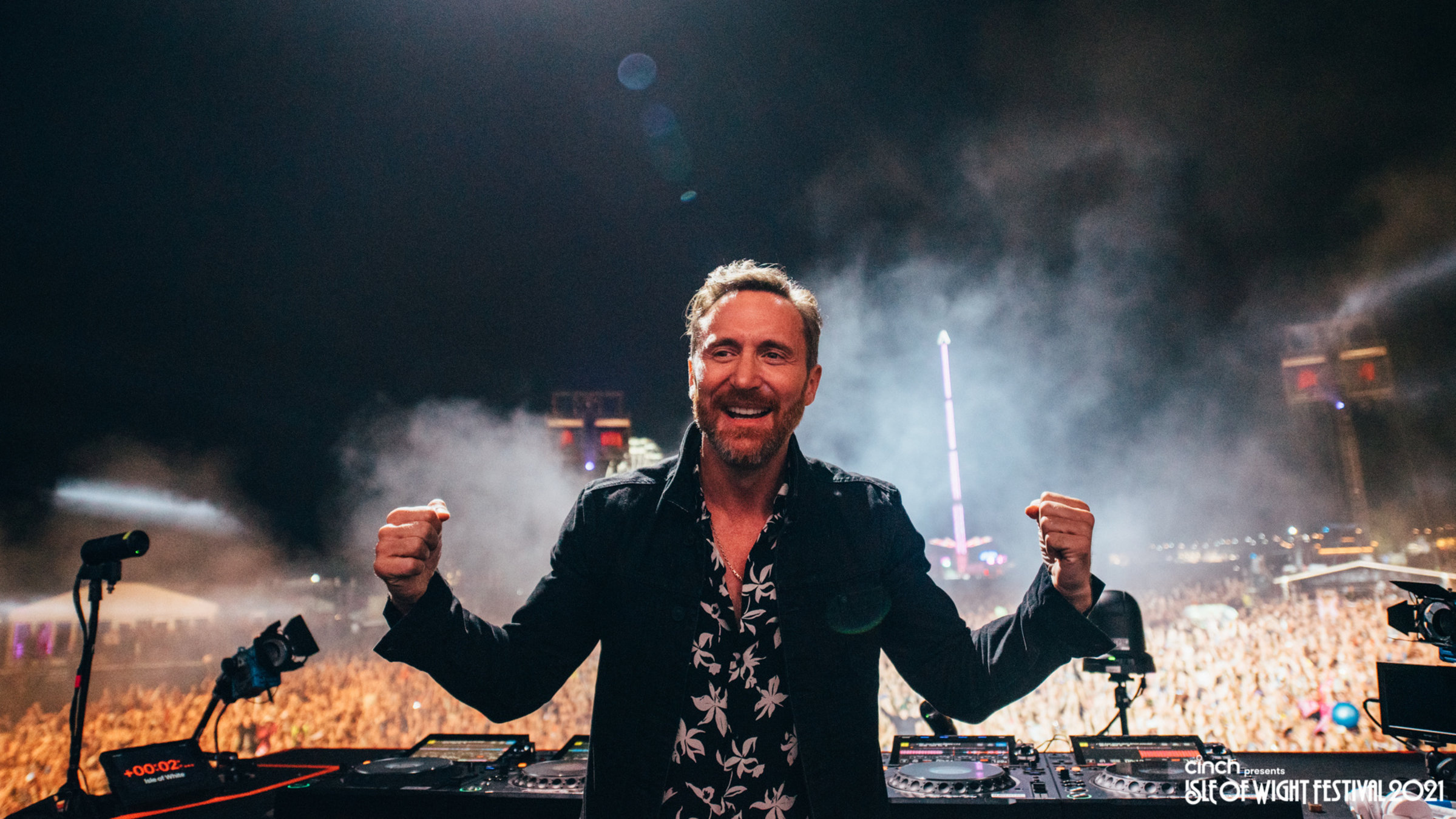"Isle of Wight Festival 2021": David Guetta steht auf der Bühne mit dem Rücken zum Publikum welches im Hintergrund zu sehen ist und lacht mit seitlich geballten Fäusten in die Kamera.