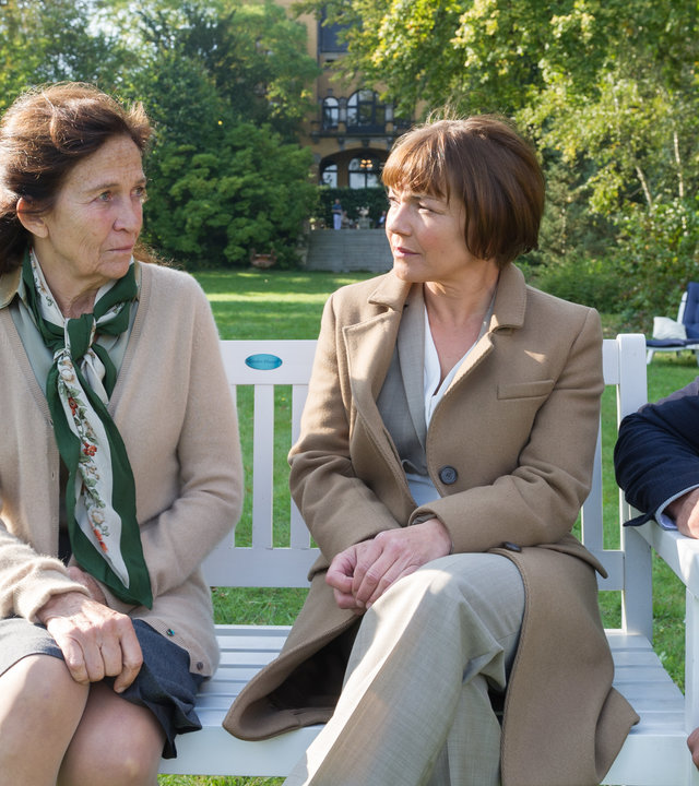 "SOKO Wismar - Goldener Herbst": Petra Kroll (Tatja Seibt) und Katrin Börensen (Claudia Schmutzler) sitzen nebeneinander auf einer weißen Gartenbank. Lars Pöhlmann (Dominic Boeer) kniet daneben. Im Hintergrund sind ein großer Garten und ein Haus zu sehen.