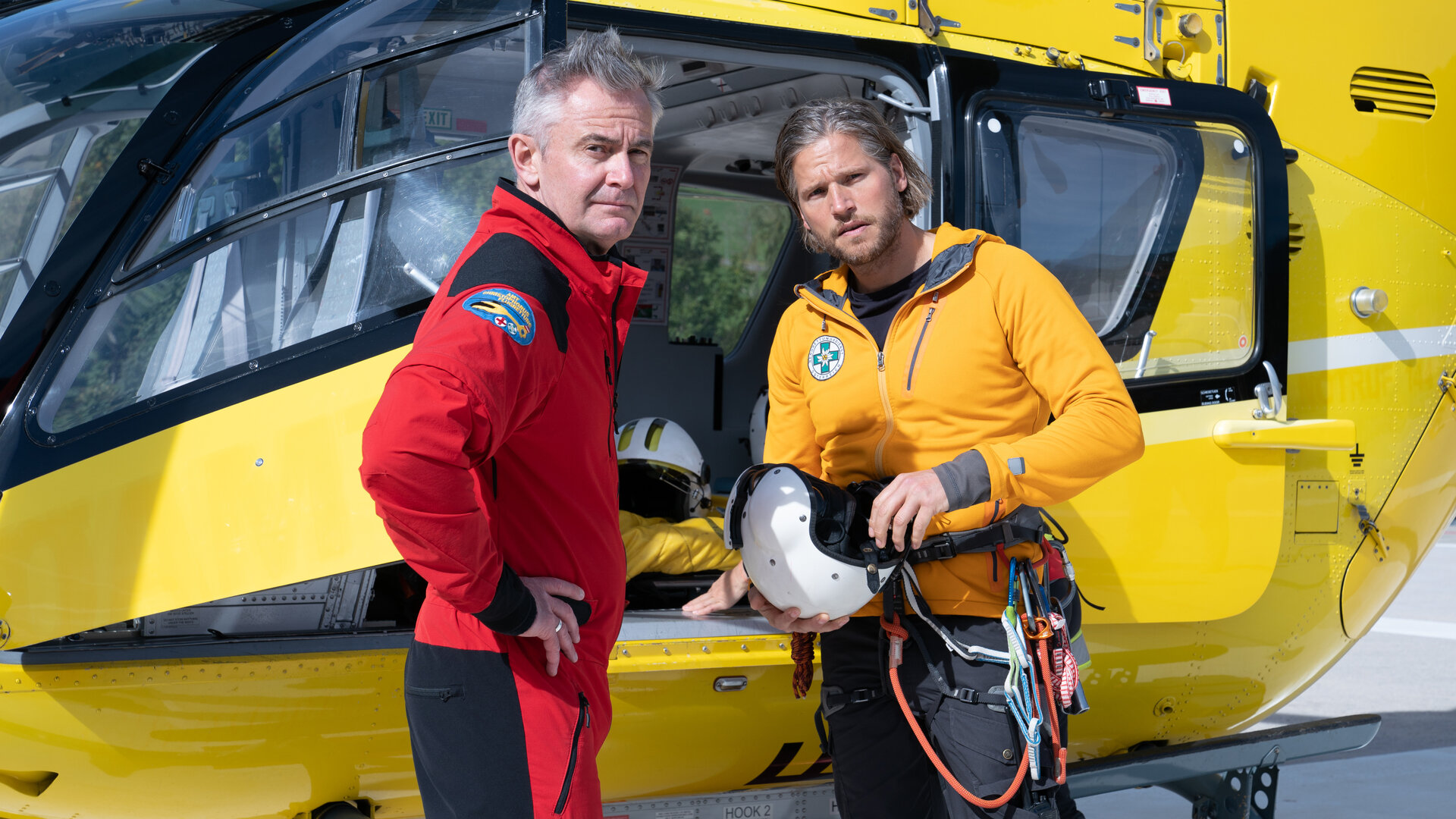 "Die Bergretter: Augen auf (2)": Vor dem Rettungshubschrauber stehen Markus (Sebastian Ströbel) und Michi (Robert Lohr) nebeneinander und blicken nachdenklich in die Ferne.