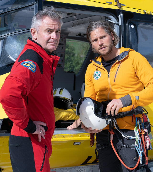 "Die Bergretter: Augen auf (2)": Vor dem Rettungshubschrauber stehen Markus (Sebastian Ströbel) und Michi (Robert Lohr) nebeneinander und blicken nachdenklich in die Ferne.