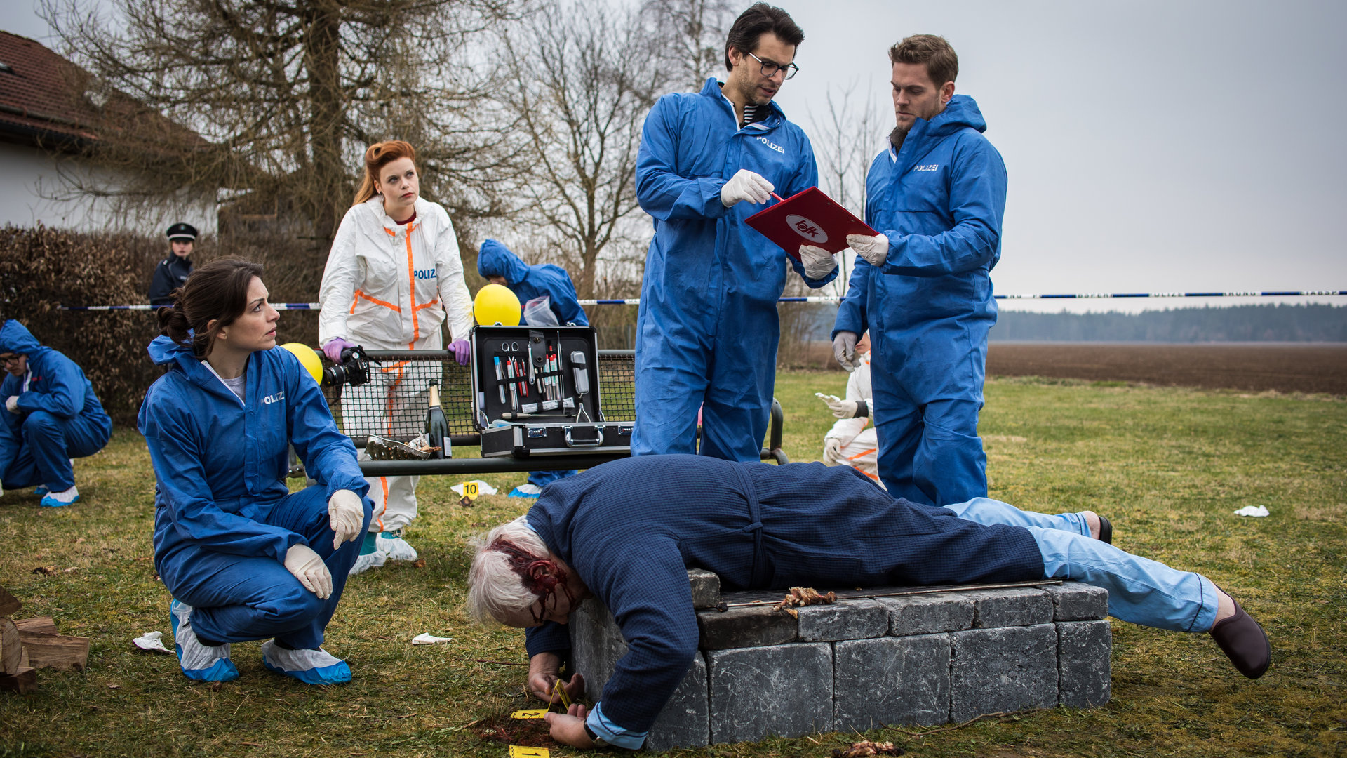 "SOKO München - Der Querulant": Am Tatort steht ein feststehender Grill aus Stein auf einer Wiese, auf ihm liegt eine Leiche. Dr. Weissenböck (Florian Odendahl) und Dominik Morgenstern (Joscha Kiefer) stehen vor der Leiche und schauen sich Daten in einer Akte näher an, Katharina Hahn (Bianca Hein) sitzt in der Hocke vor der Leiche. Im Hintergrund ist eine Parkbank zu sehen, ein Koffer von der Spusi wurde dort abgestellt. Billie Curio (Sina Reiß) steht hinter der Bank und stützt sich auf der Rückenlehne ab. Sie hat eine Fotokamera in ihrer rechten Hand.