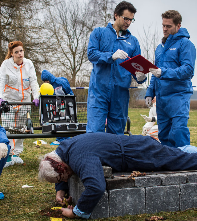 "SOKO München - Der Querulant": Am Tatort steht ein feststehender Grill aus Stein auf einer Wiese, auf ihm liegt eine Leiche. Dr. Weissenböck (Florian Odendahl) und Dominik Morgenstern (Joscha Kiefer) stehen vor der Leiche und schauen sich Daten in einer Akte näher an, Katharina Hahn (Bianca Hein) sitzt in der Hocke vor der Leiche. Im Hintergrund ist eine Parkbank zu sehen, ein Koffer von der Spusi wurde dort abgestellt. Billie Curio (Sina Reiß) steht hinter der Bank und stützt sich auf der Rückenlehne ab. Sie hat eine Fotokamera in ihrer rechten Hand.