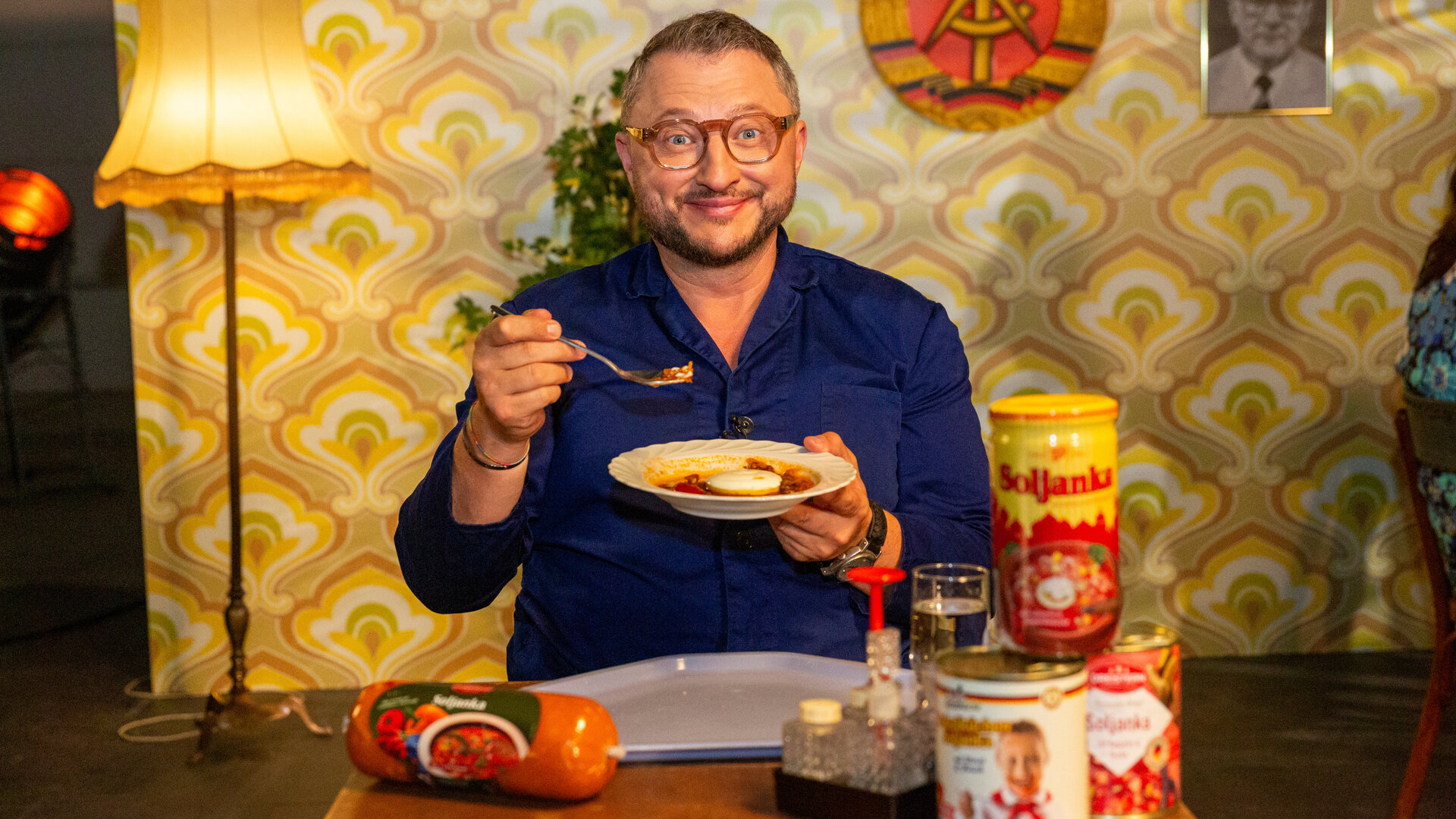 "besseresser: Die Tricks in DDR-Produkten - Sebastian Lege deckt auf": Sebastian Lege hält einen Teller mit Soljanka in der linken Hand, während er in der rechten Hand einen Löffel hält. Vor ihm stehen verschiedene Ost-Produkte auf dem Tisch.