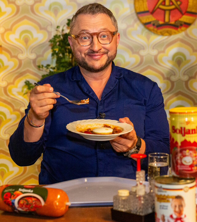 "besseresser: Die Tricks in DDR-Produkten - Sebastian Lege deckt auf": Sebastian Lege hält einen Teller mit Soljanka in der linken Hand, während er in der rechten Hand einen Löffel hält. Vor ihm stehen verschiedene Ost-Produkte auf dem Tisch.