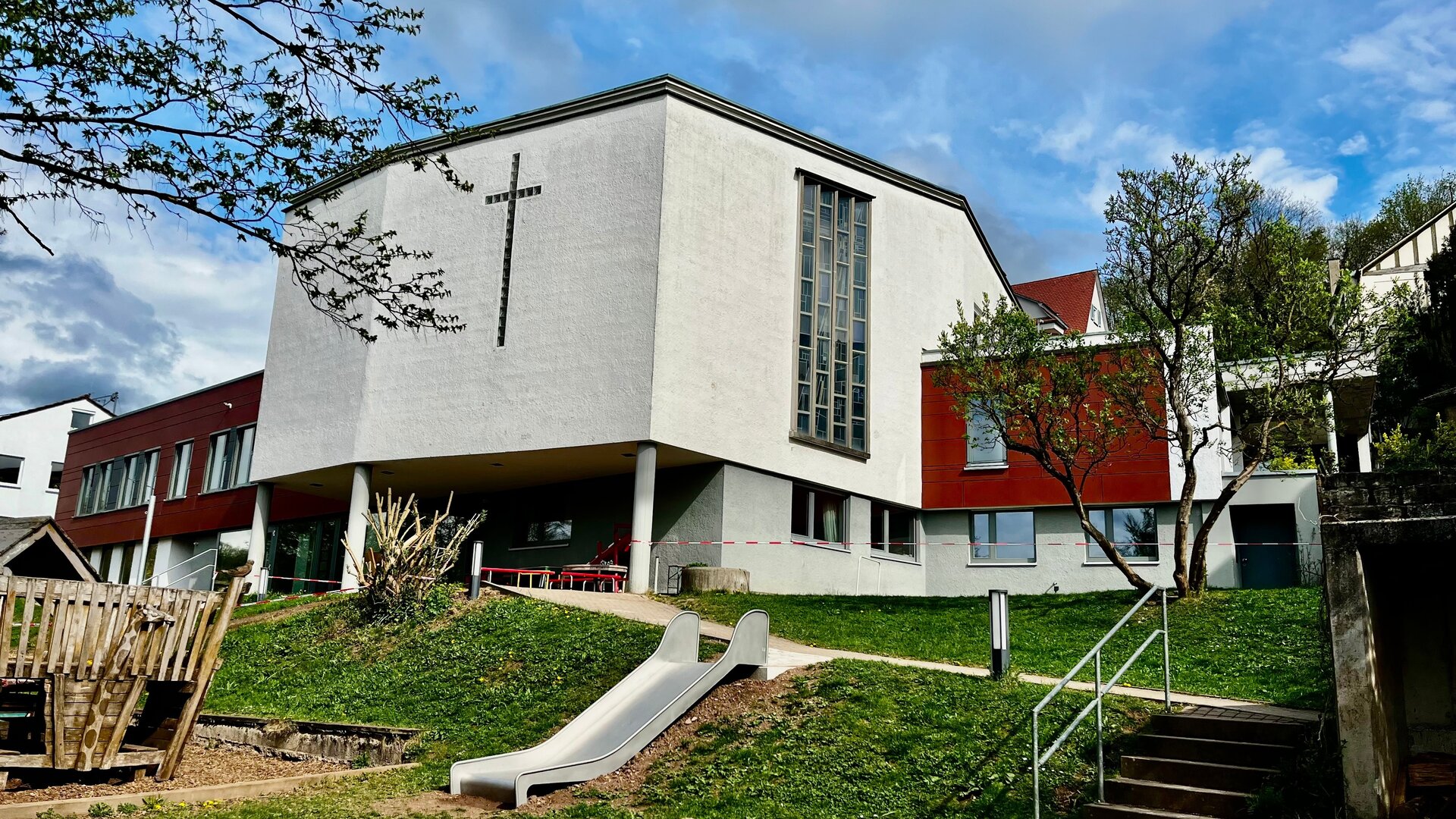 "Evangelischer Gottesdienst - Hoffnungsgroß statt hoffnungslos": Außenansicht der 1961 erbauten Kreuzkirche Tübingen: ein moderner Bau ohne Kirchturm, dafür mit einem großen, silbergrauen Kreuz an der Frontseite der Kirche. Im Vordergrund befindet sich der  Garten und Spielplatz der angrenzenden Kindertagesstätte.