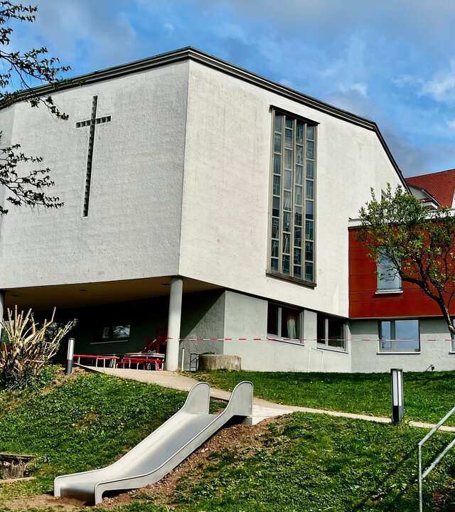 "Evangelischer Gottesdienst - Hoffnungsgroß statt hoffnungslos": Außenansicht der 1961 erbauten Kreuzkirche Tübingen: ein moderner Bau ohne Kirchturm, dafür mit einem großen, silbergrauen Kreuz an der Frontseite der Kirche. Im Vordergrund befindet sich der  Garten und Spielplatz der angrenzenden Kindertagesstätte.
