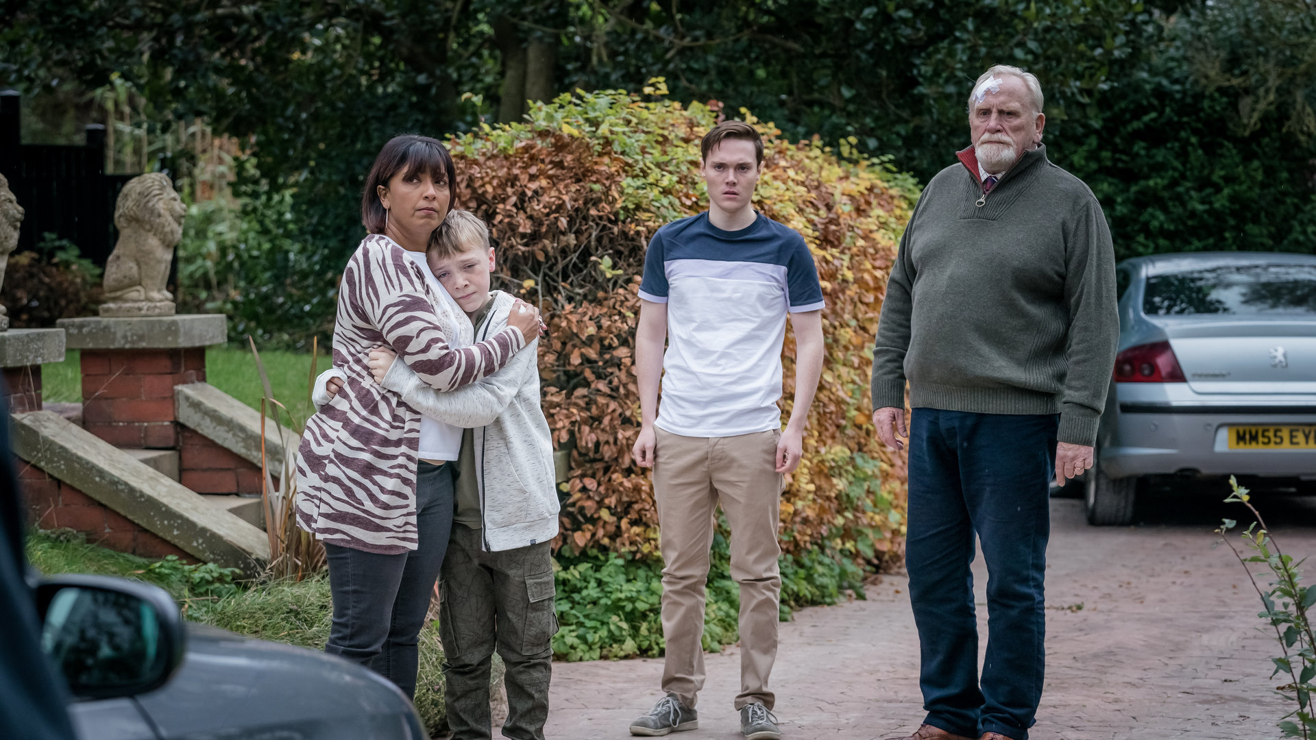 "The Bay - Verbundenheit": Außenaufnahme vor einem Haus. Stella Bradwell (Sunetra Sarker) umarmt Oliver Marshbrook (Leo Ashton). Jamie Marshbrook (Jack Archer) und Bill Bradwell (James Cosmo) stehen daneben. Alle blicken schockiert in dieselbe Richtung.