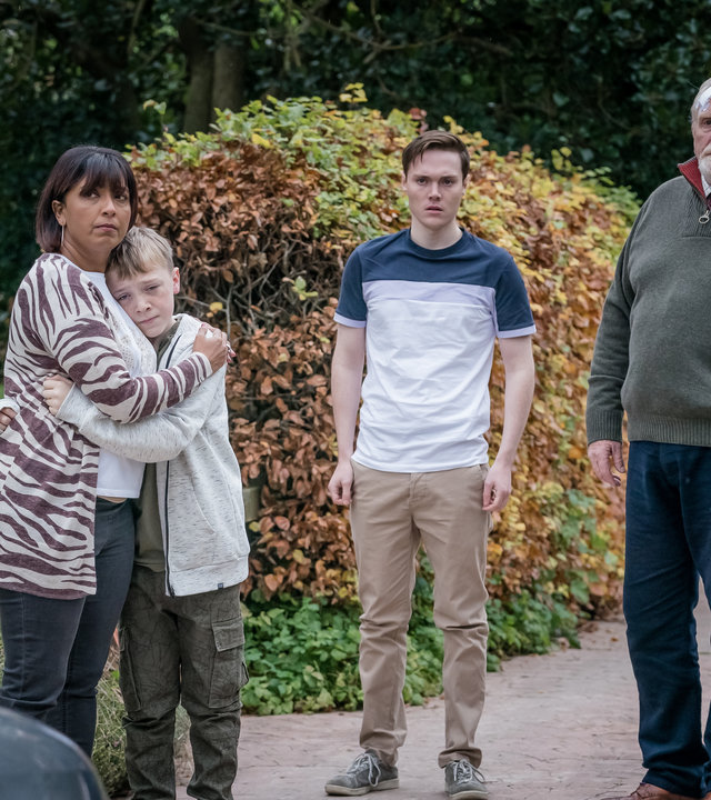 "The Bay - Verbundenheit": Außenaufnahme vor einem Haus. Stella Bradwell (Sunetra Sarker) umarmt Oliver Marshbrook (Leo Ashton). Jamie Marshbrook (Jack Archer) und Bill Bradwell (James Cosmo) stehen daneben. Alle blicken schockiert in dieselbe Richtung.