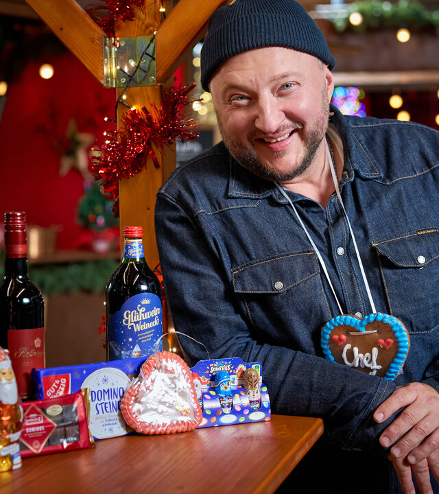 "besseresser: Die Tricks in Glühwein, Lebkuchen & Co. - Sebastian Lege deckt auf": Produktentwickler Sebastian Lege steht an einem Weihnachtsmarkt-Stand, um seinen Hals trägt er ein Lebkuchenherz mit der Aufschrift "Chef", neben ihm auf dem Tisch sind Glühweinflaschen, Schoko-Nikoläuse und Dominosteine angeordnet.