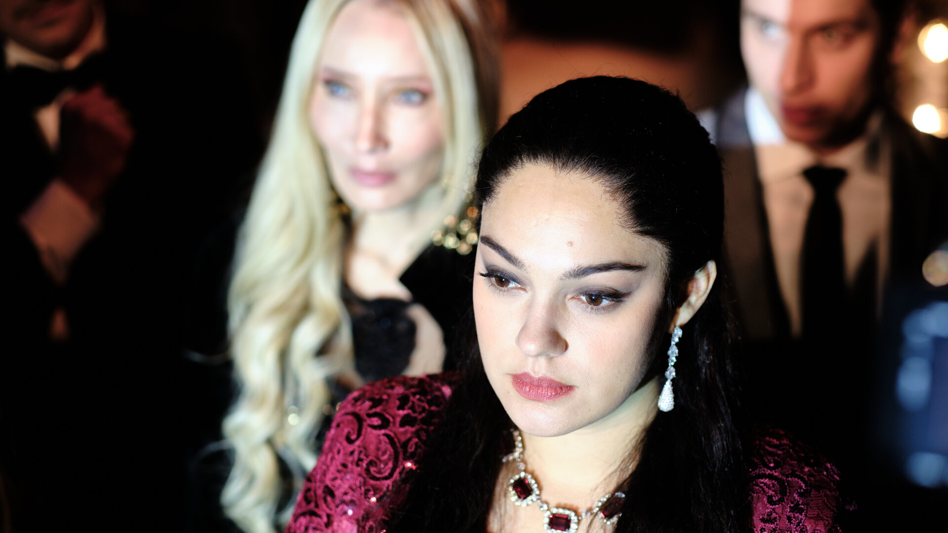 "Take the Money and Run - One Dream": Ruja Ignatova (Nilam Farooq) steht in einem glitzernden Kleid und auffälligem Schmuck im Vordergrund, ihr Blick ist konzentriert nach vorne gerichtet. Hinter ihr stehen Elva Gunnarsdottir (Tina Balthazar) und Jonas Kurth (László Branko Breiding).