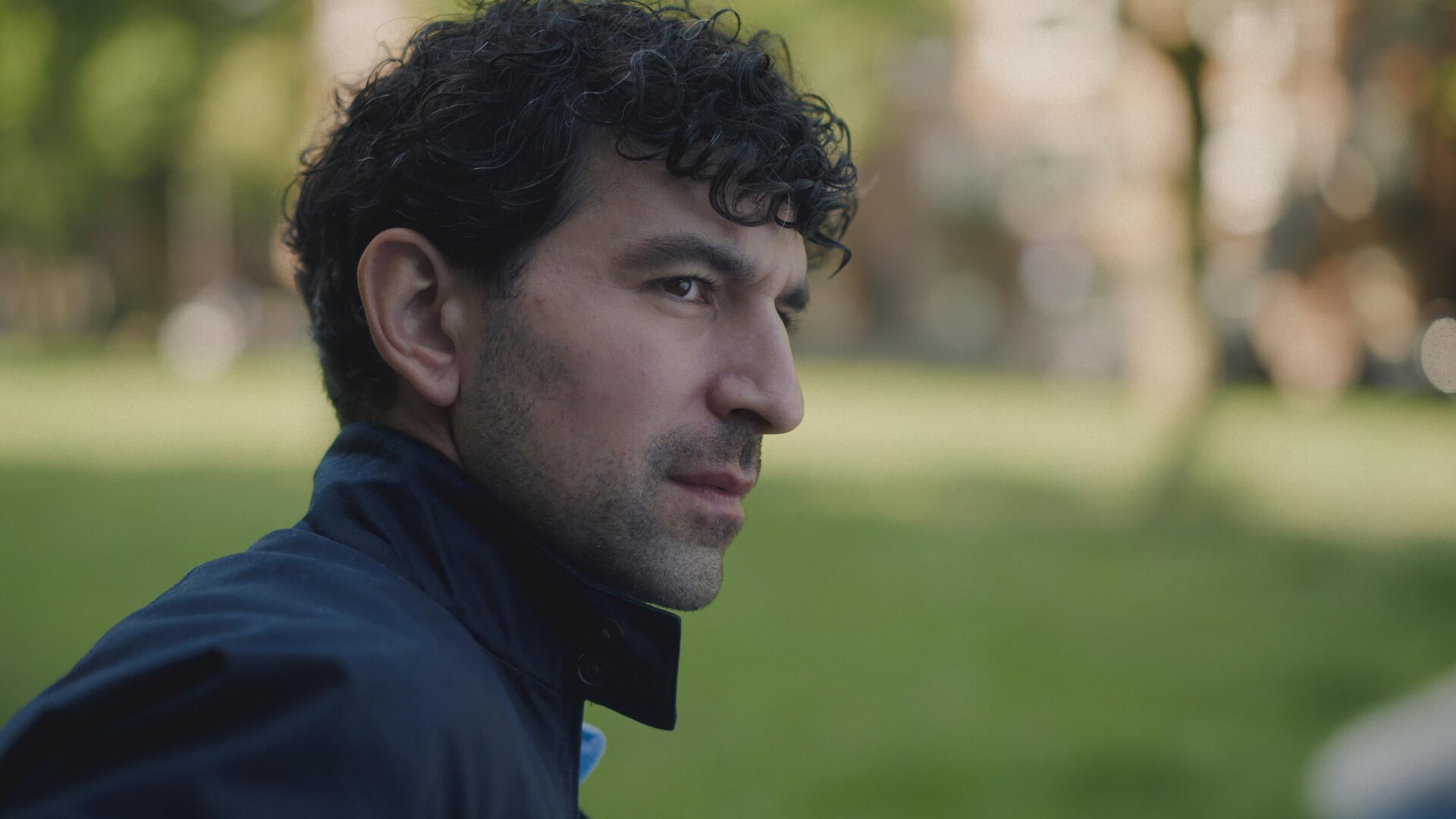 "Blind Sherlock - Miese Ratte": Yaqub (Sinan Eroglu) steht im Grünen und blickt zur Seite. Er macht einen skeptischen Eindruck.